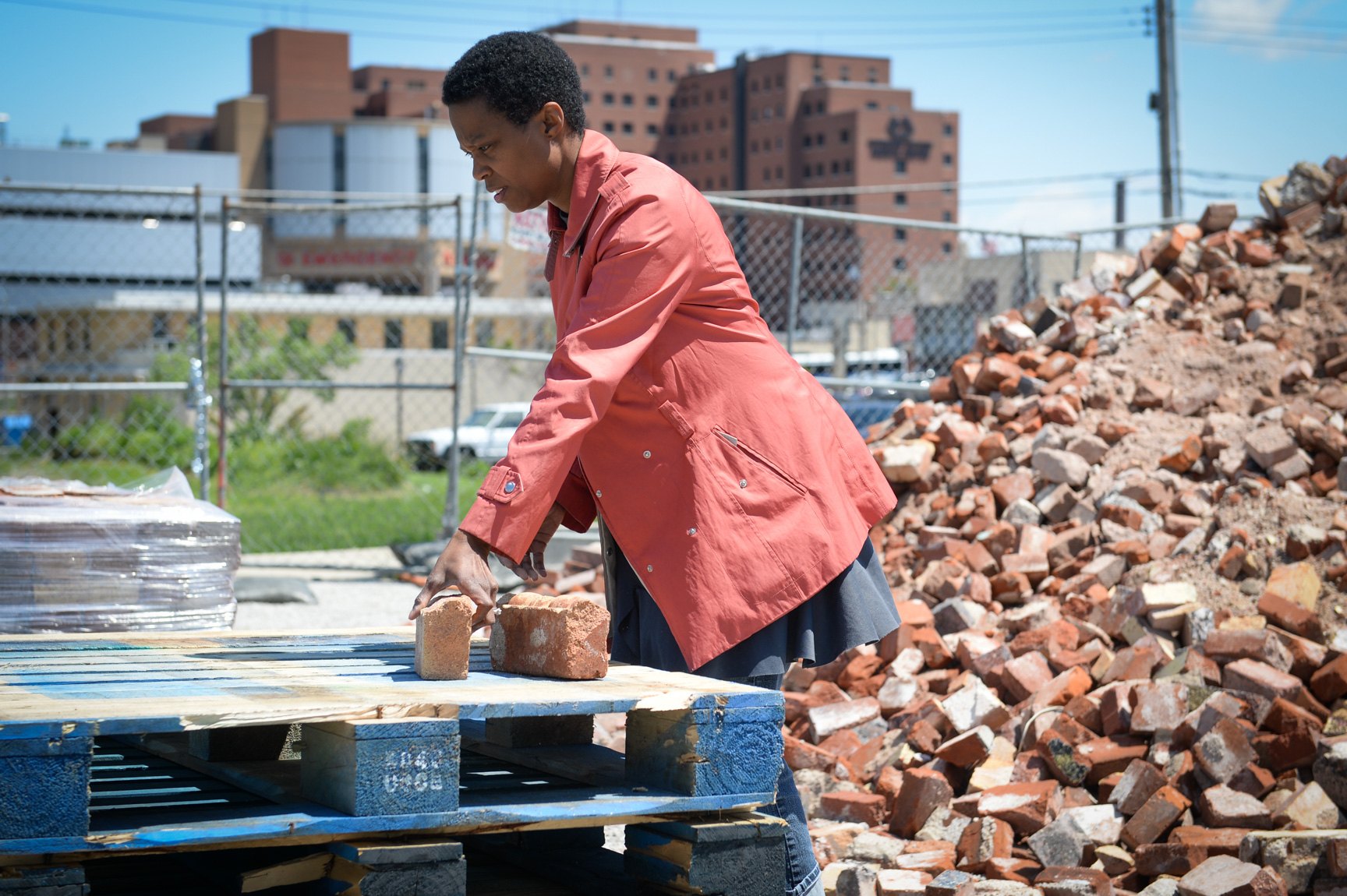Amanda Williams places a brick on a stack of blue pallets with a large heap of bricks in the background.