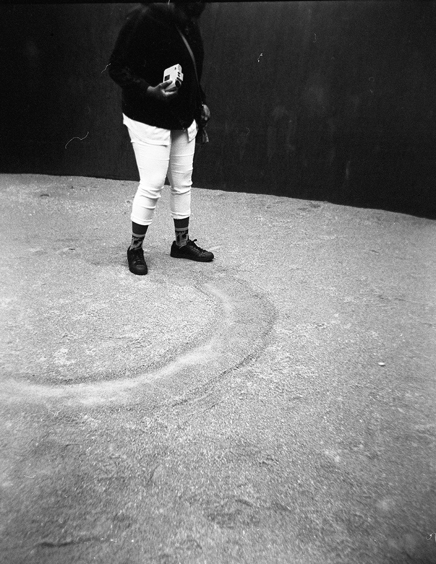 A black and white shot of a participant standing in "Joe" by Richard Serra, holding a camera.