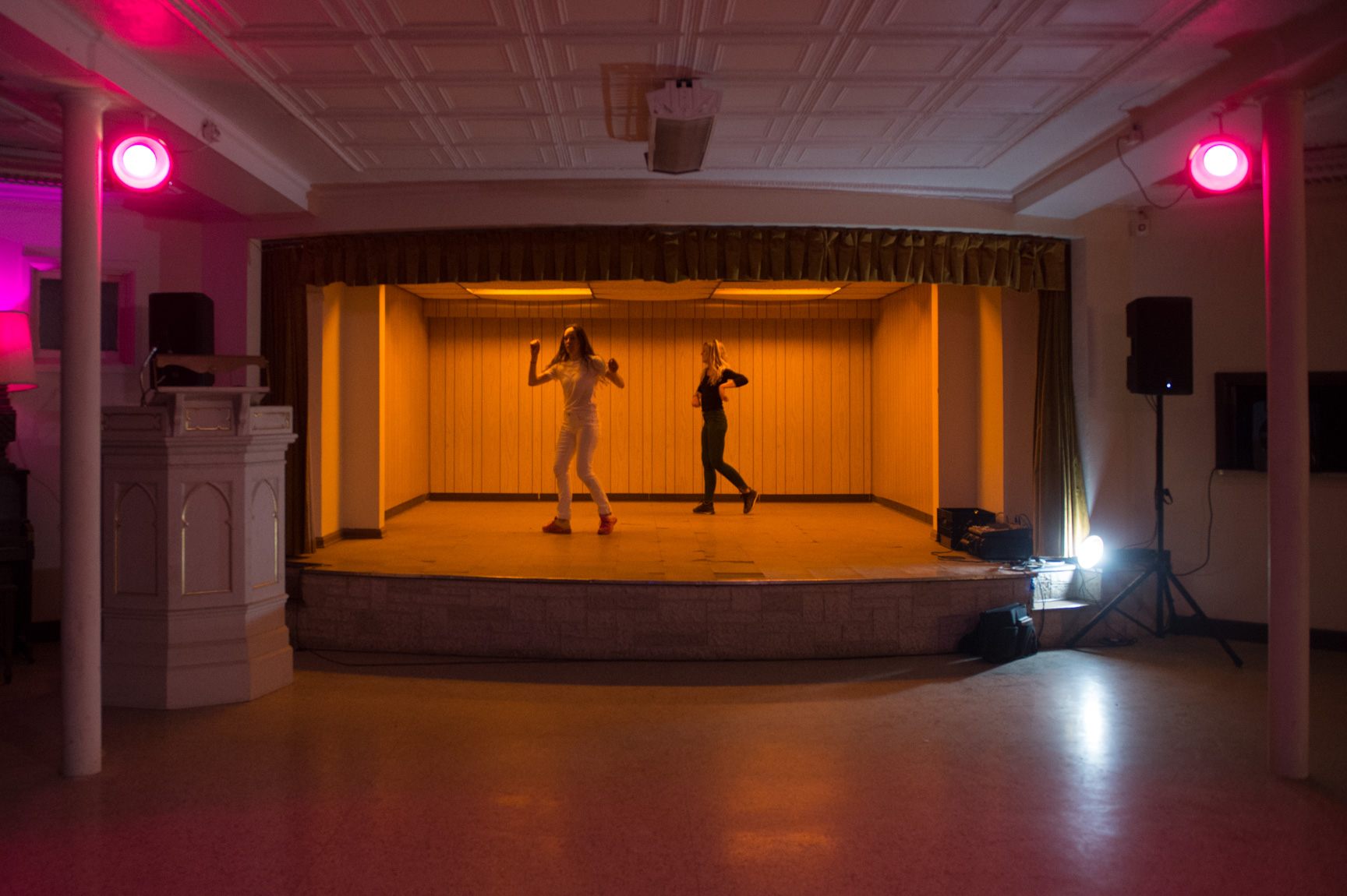 Two AUNTS dancers performs on a small stage flanked by two purple stage lights in the former St. Matthew’s United Church of Christ.