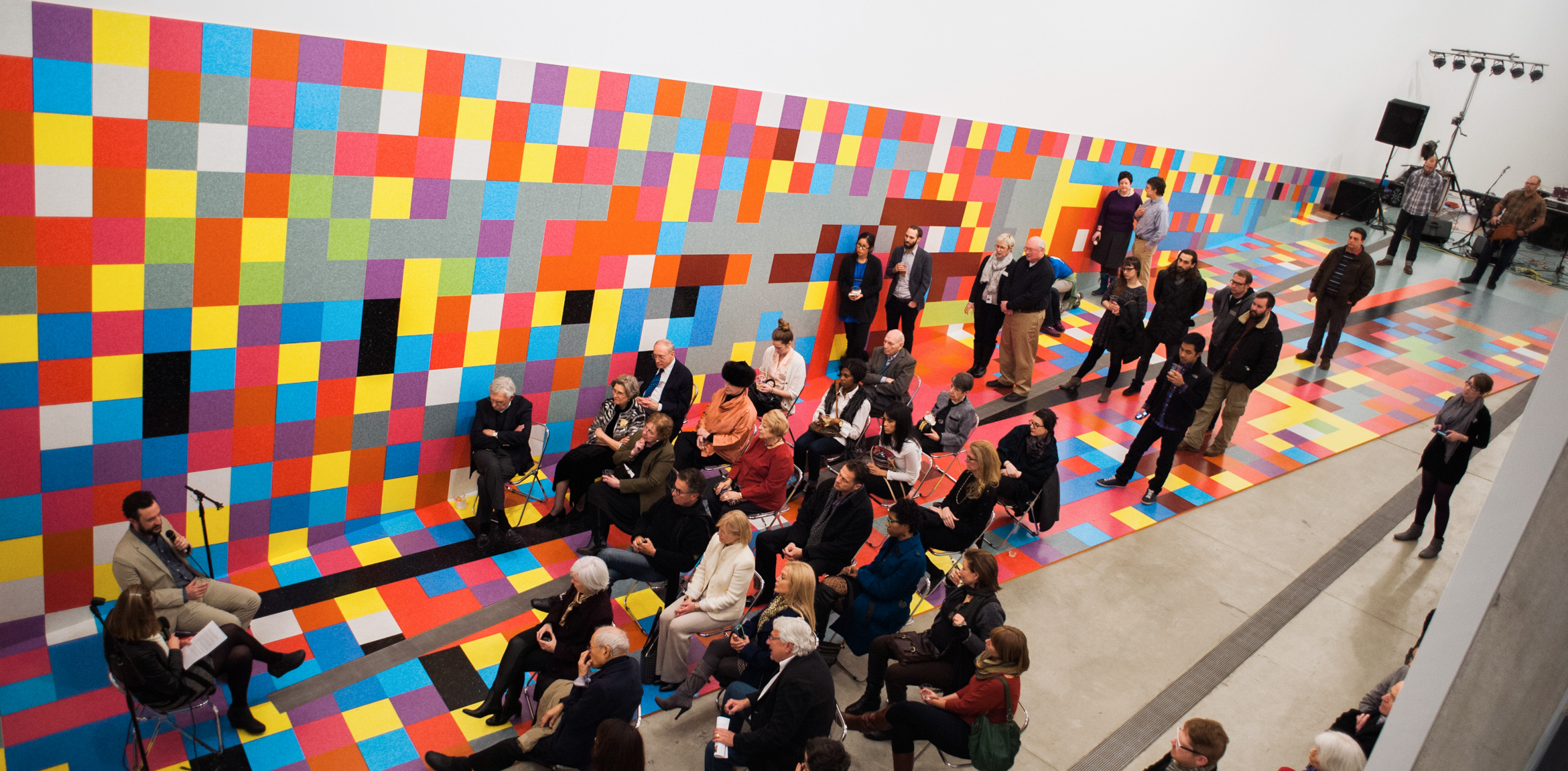 David Scanavino and an interviewer sit and speak into microphones for a seated audience in the main gallery, on Scanavino's colorful gridded installation.