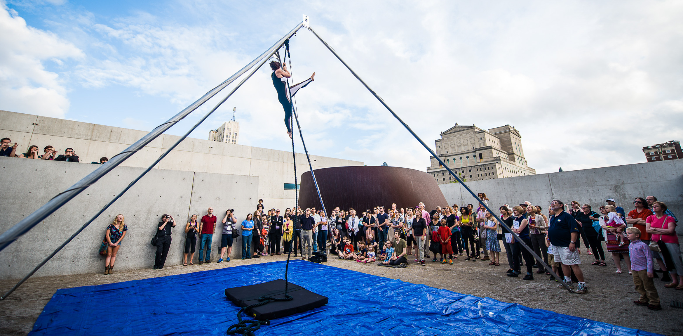 A trapeze artist from Circus Flora climbs a rope in a constructed pyramid, performing for large crowd in the Courtyard.