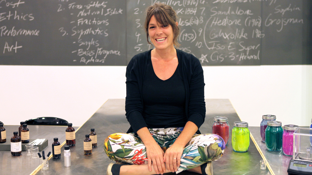 The Institute for Art and Olfaction Founder and Director Saskia Wilson Brown sits cross-legged on a metal desk with experimental supplies and smiles for the camera in front of a blackboard.