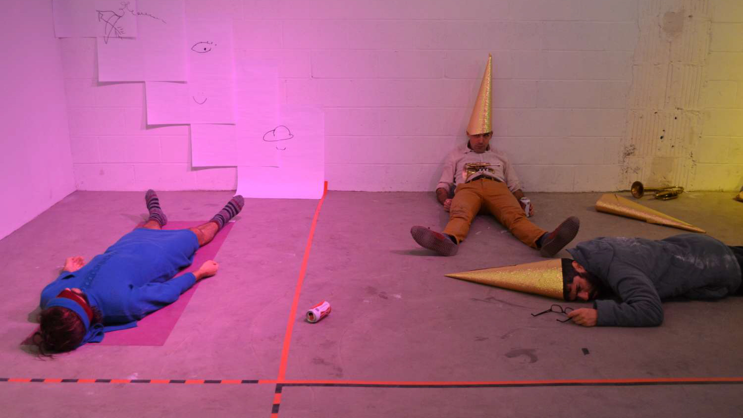 Three AUNTS performers lie on the ground. Two wear gold cones on their heads and one wears a blue dress and a blindfold.