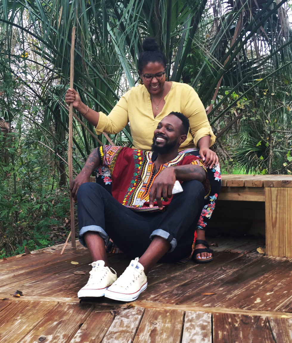 Artists Danielle and Kevin McCoy laugh together outdoors. Danielle sits on a wood bench and Kevin sits on the porch floor before her.