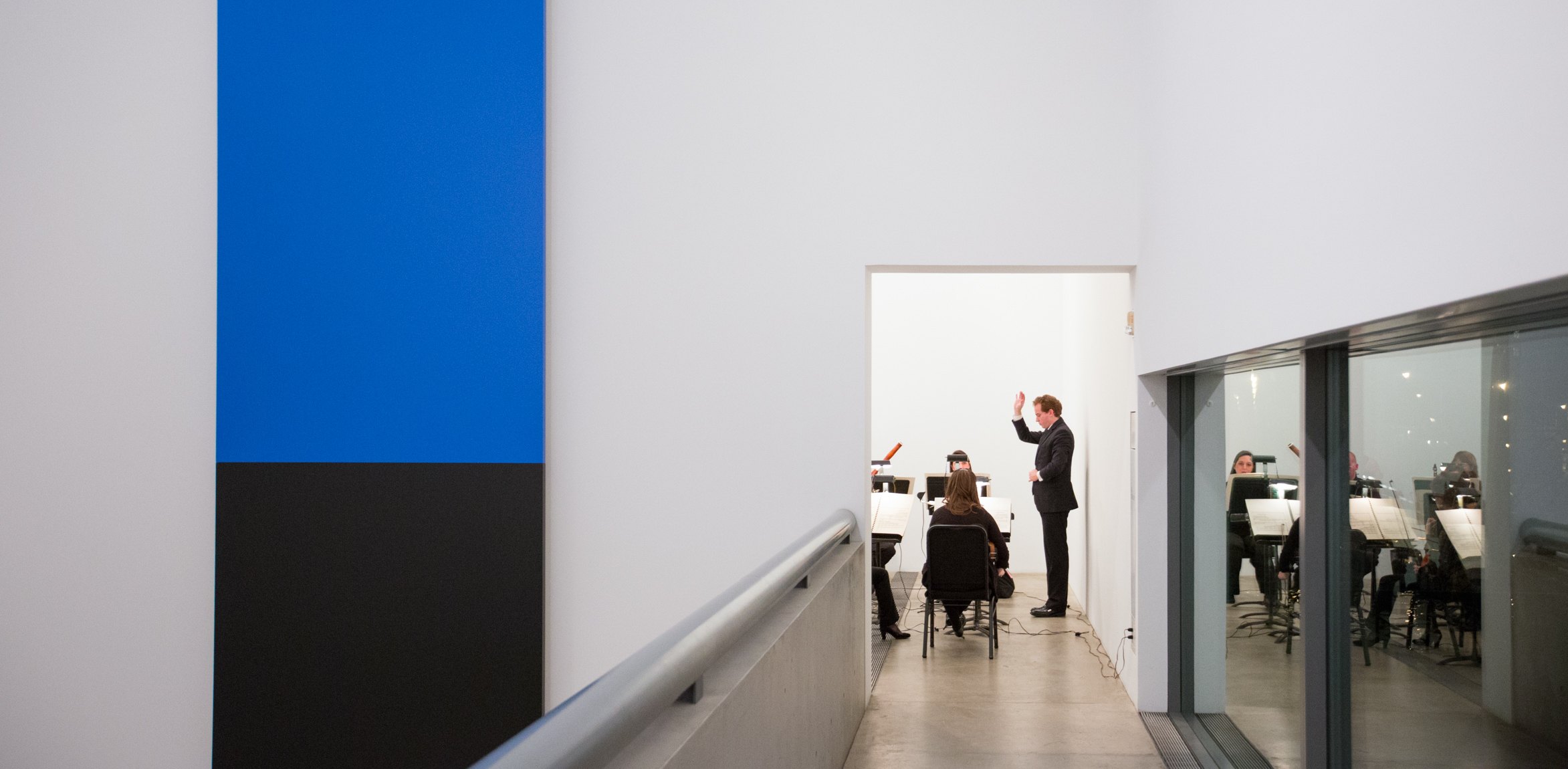 A view from the Main Gallery corridor of a conductor and symphony performing in the Cube Gallery. To the left is Ellsworth Kelly's 