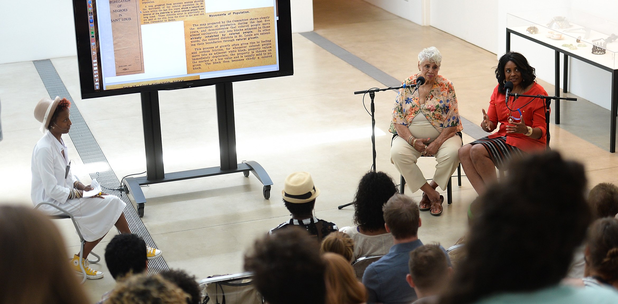 Activist Tara Tee sits to the left of a large monitor in the Lower Main Gallery, and two participants sit to the right and speak into microphones to the audience on the Main Staircase.