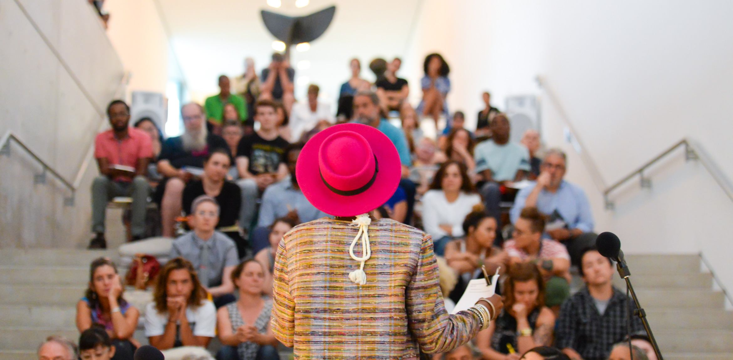 A view from behind of Tara Tee, wearing a bright pink hat, speaking to an audience on the Main Staircase.