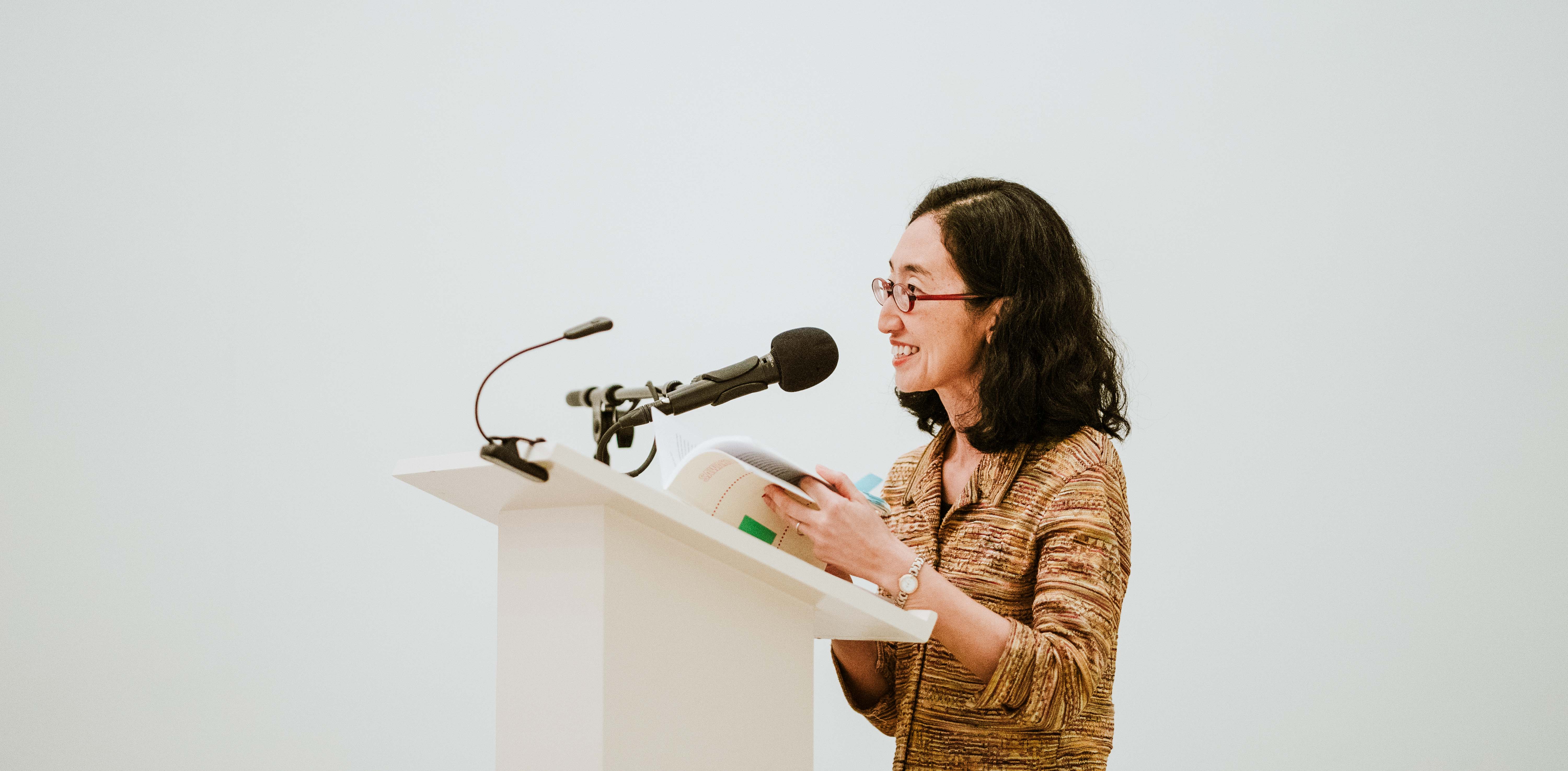 Poet Sawako Nakayasu smiles at the audience from behind a podium with a microphone.