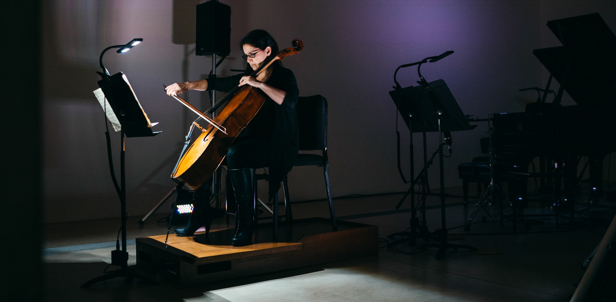 A cellist performs in a dark gallery, lit by a music stand light.