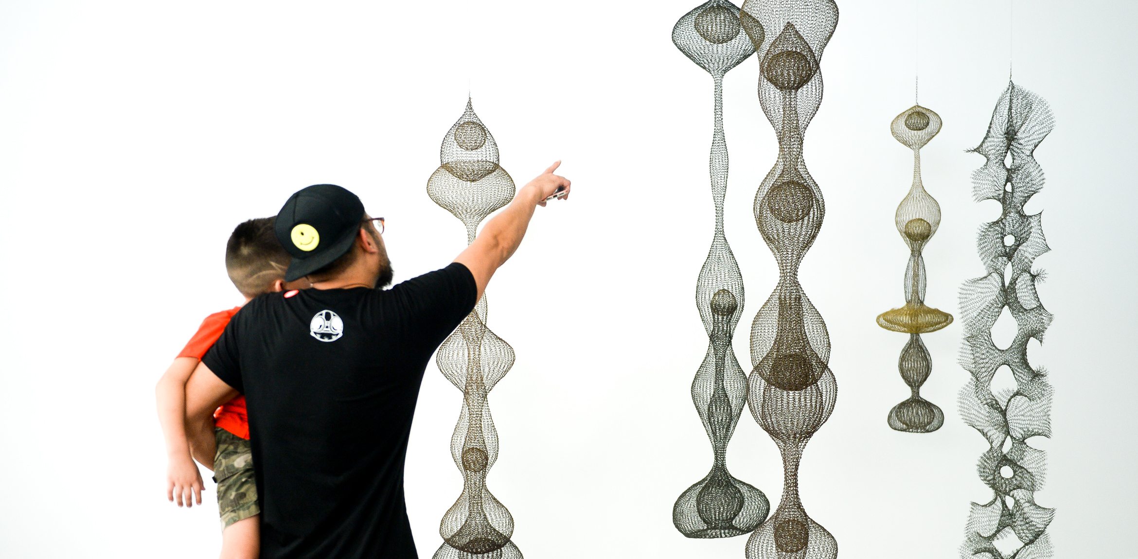 A visitor carrying a child points at a Ruth Asawa's hanging wire sculptures.