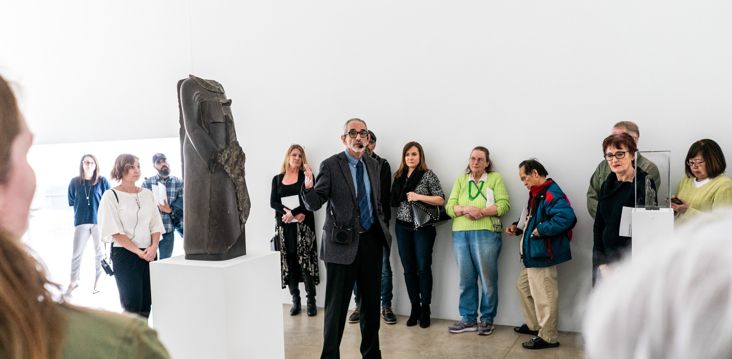 Ed Bleiberg speaks in the Main Gallery to a large circle of attendees, gesturing at a beheaded Egyptian sculpture.