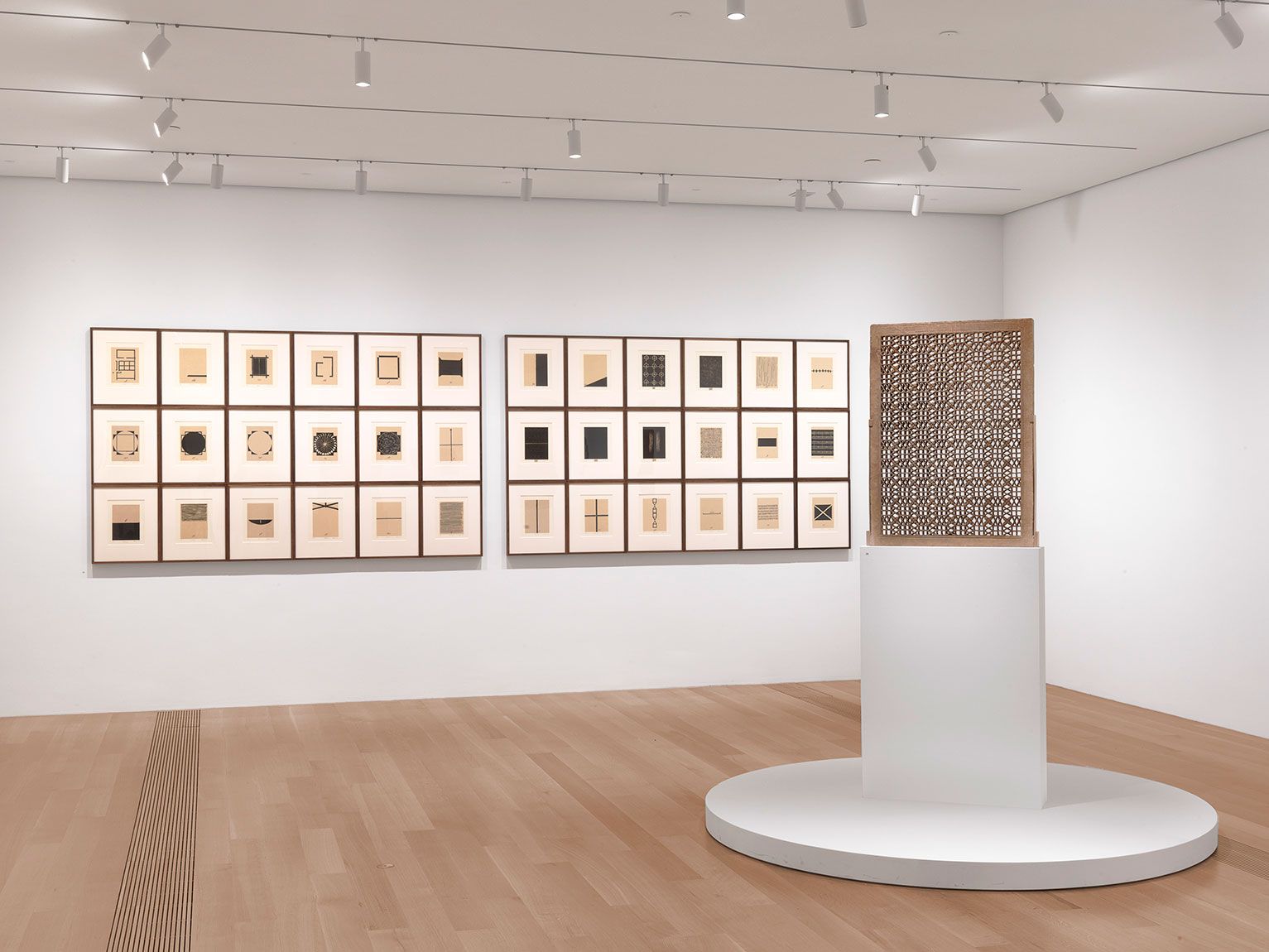 An exhibition view of the Lower West Gallery, including a portfolio by Zarina titled "Home is a Foreign Place," 36 framed woodcut prints hung in a grid on the south wall, and a red sandstone "Jali," or windowscreen, by an unidentified artist on a pedestal in the center.