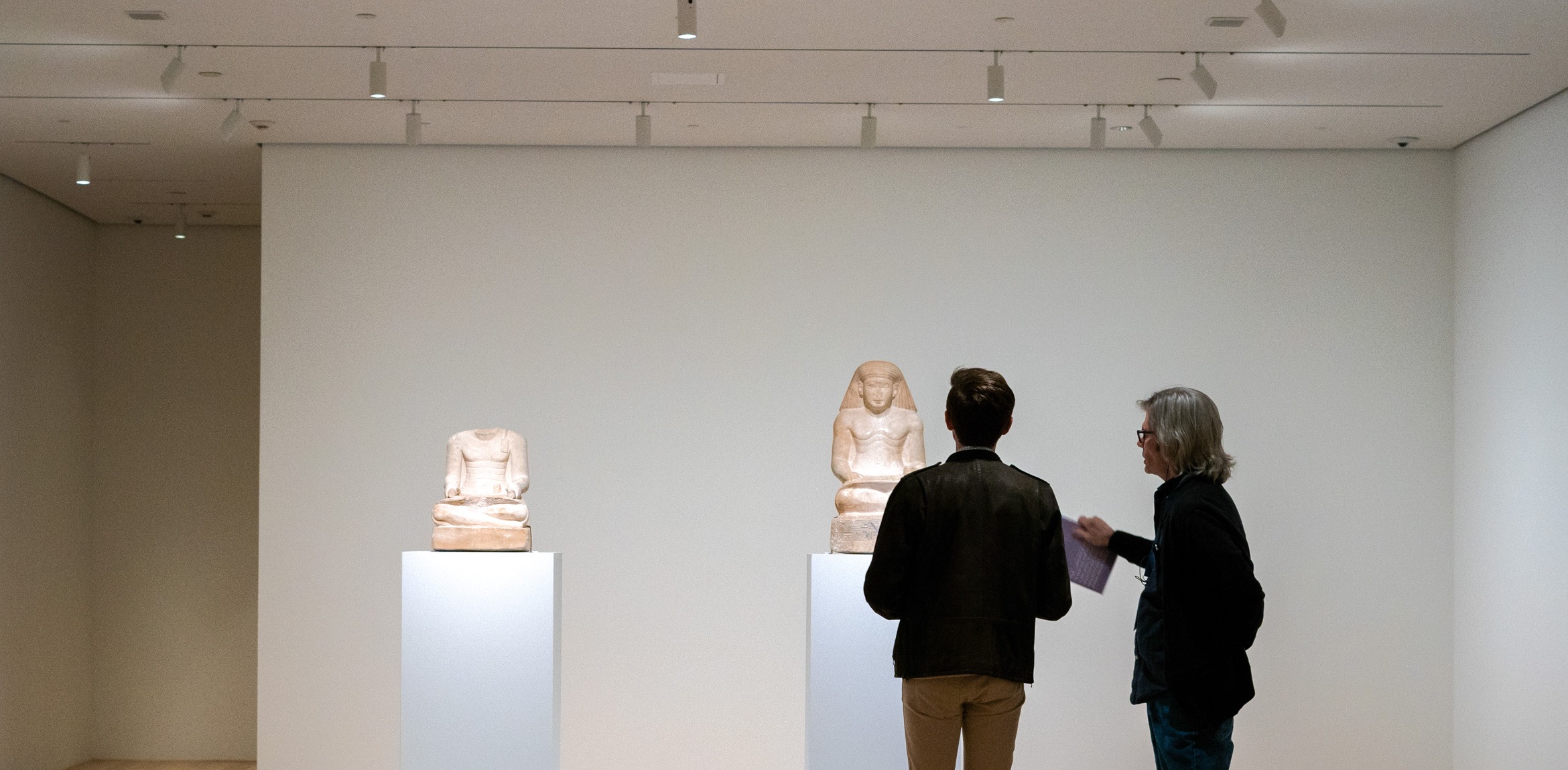 Two visitors view two white limestone sculptures in the Lower East Gallery titled 