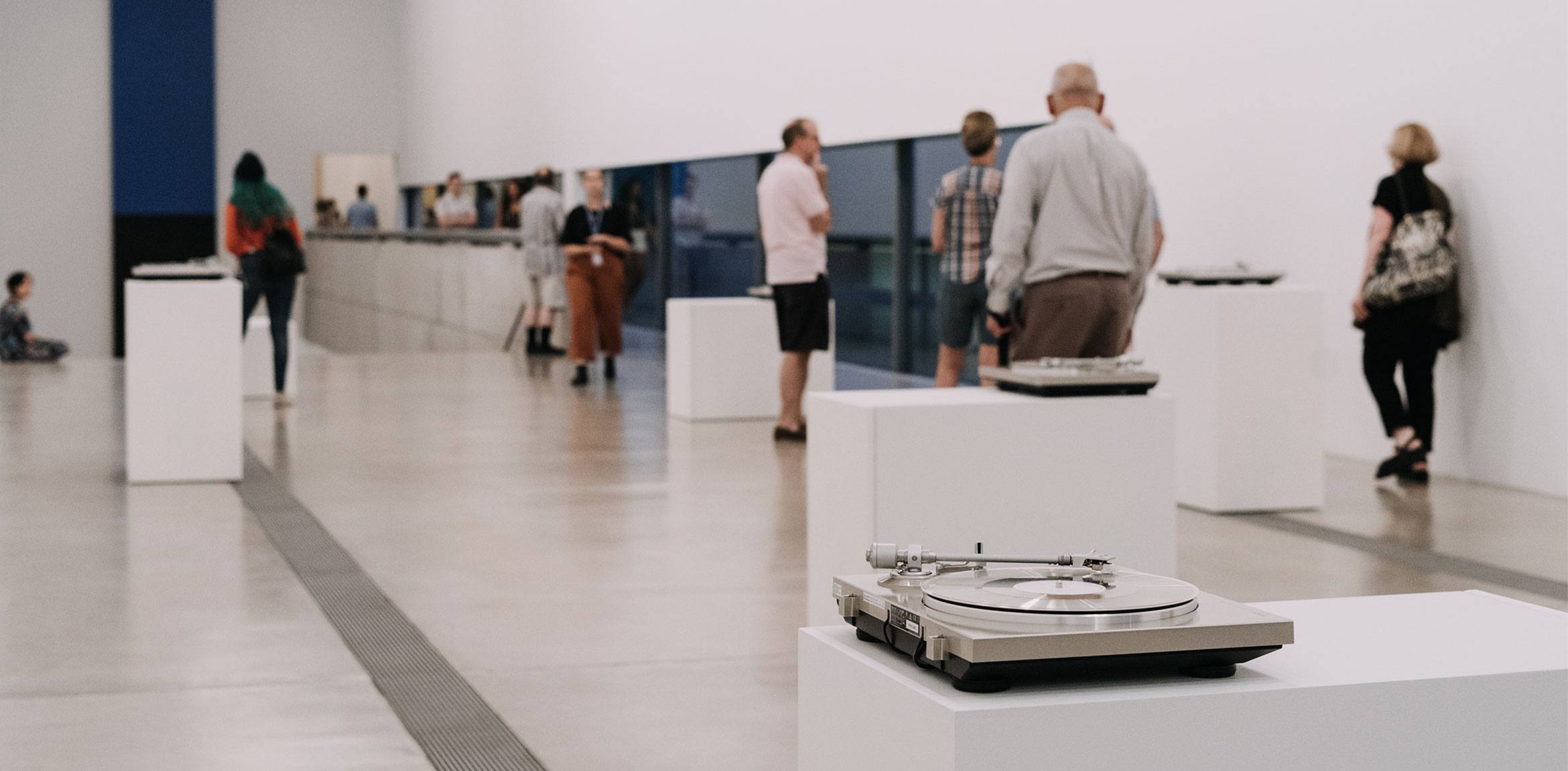 Visitors congregate in the Main Gallery, and the camera focuses on one of Susan Philipsz's turntables on a podium.