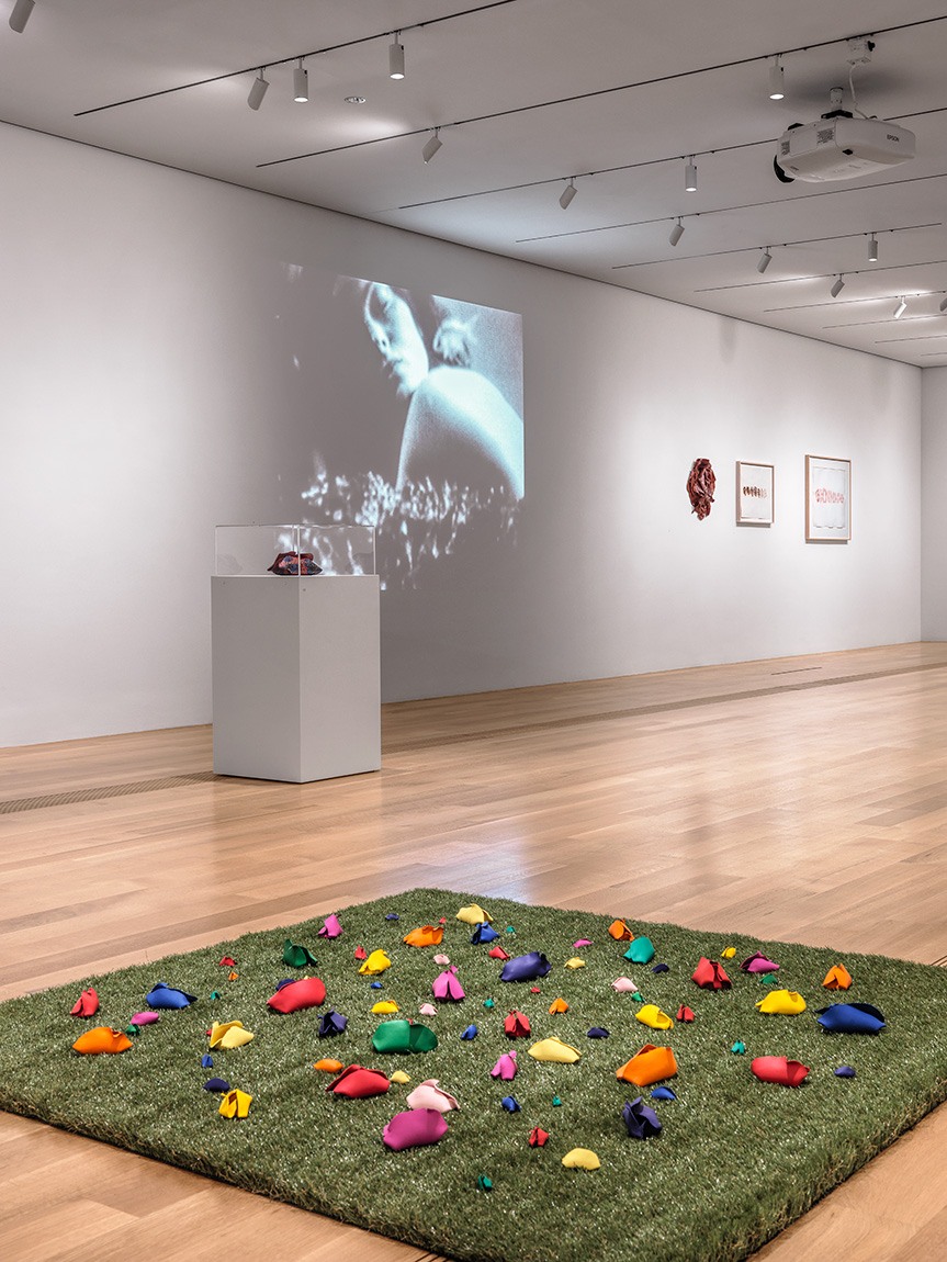 A square of green turf installed with colorful painted clay sculptures; in the background, a vitrine, projection, two framed works, and a latex wall work are visible
