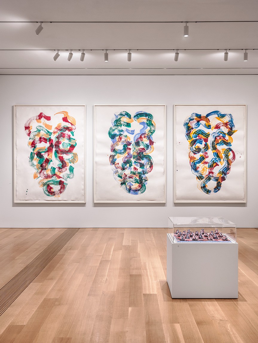 (Background) three large paintings of abstracted faces; (Foreground) a vitrine holding a number of painted clay sculptures