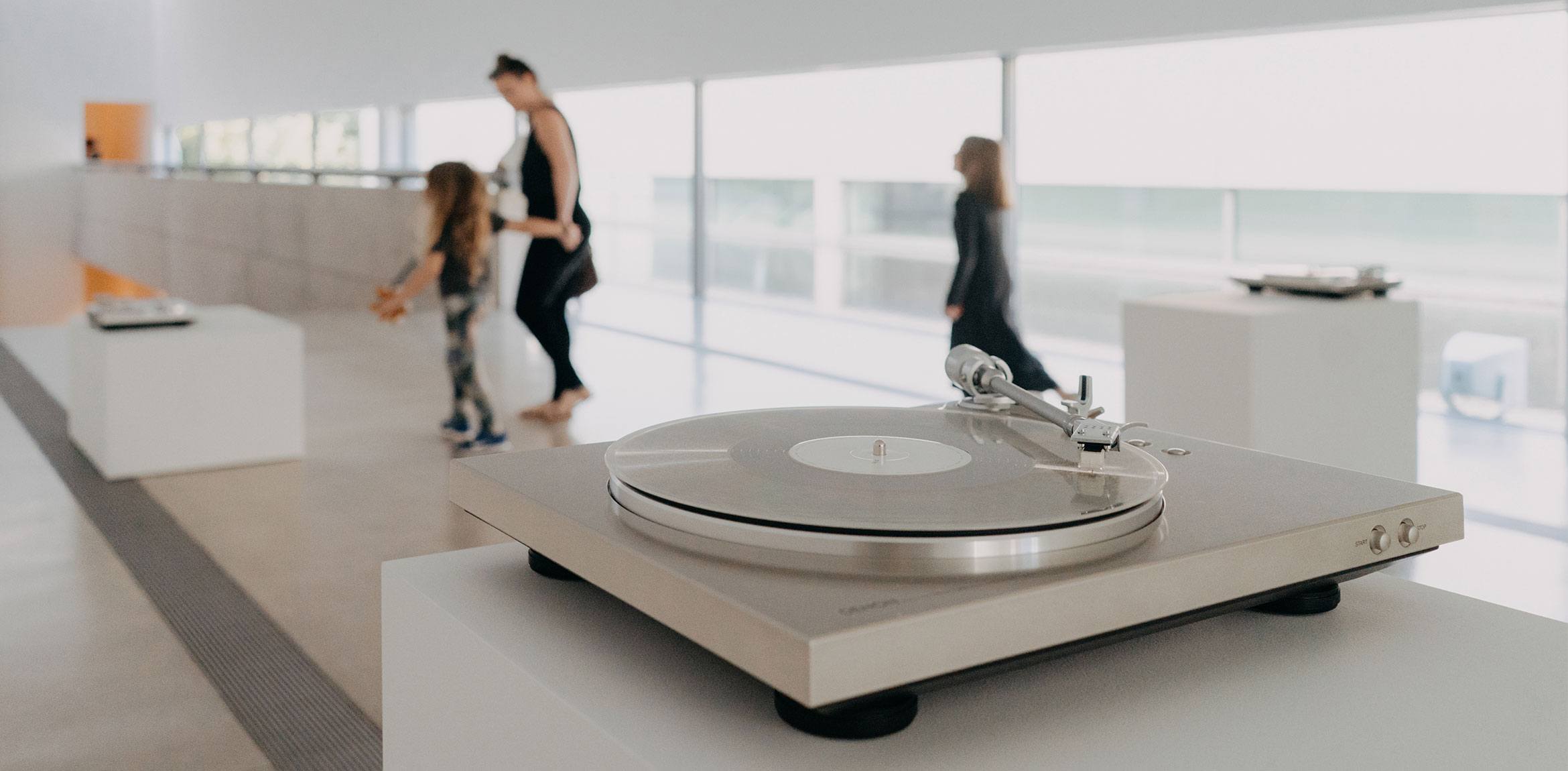 A closeup of one of Susan Philipsz's silver turntables on a pedestal in the Main Gallery. In the background, a visitor and their children walk through the exhibition.