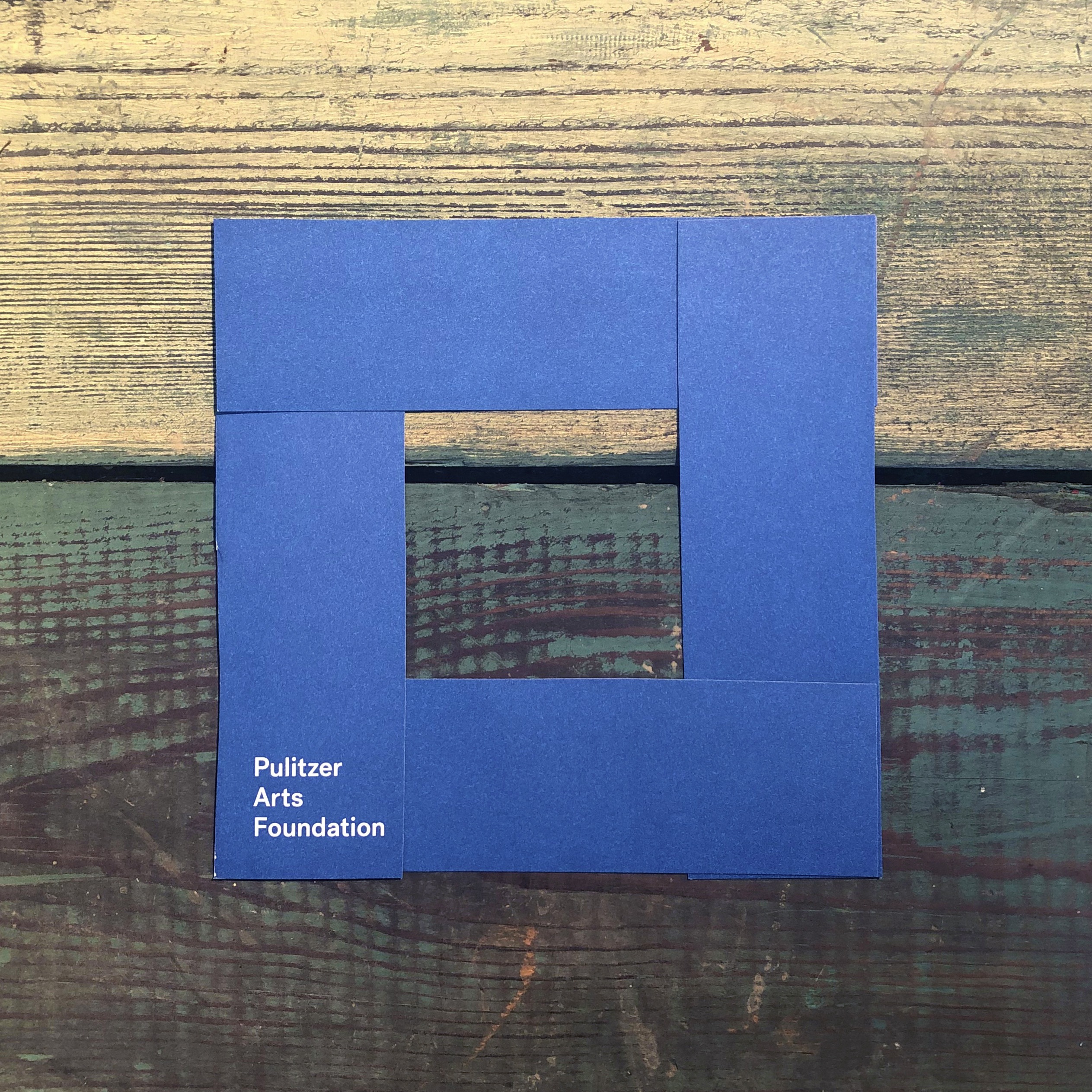 A handmade square viewfinder, made with a blue Pulitzer catalogue, on a wooden surface.