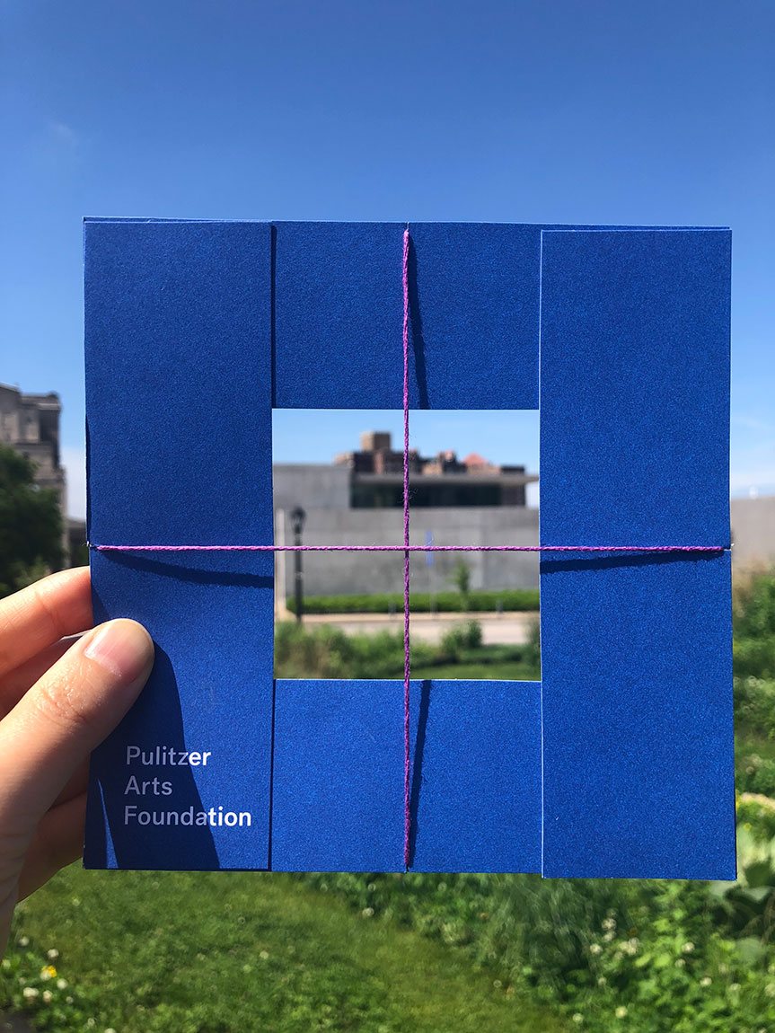 A handmade square viewfinder, made with a blue Pulitzer catalogue and string, framing the Ando building.