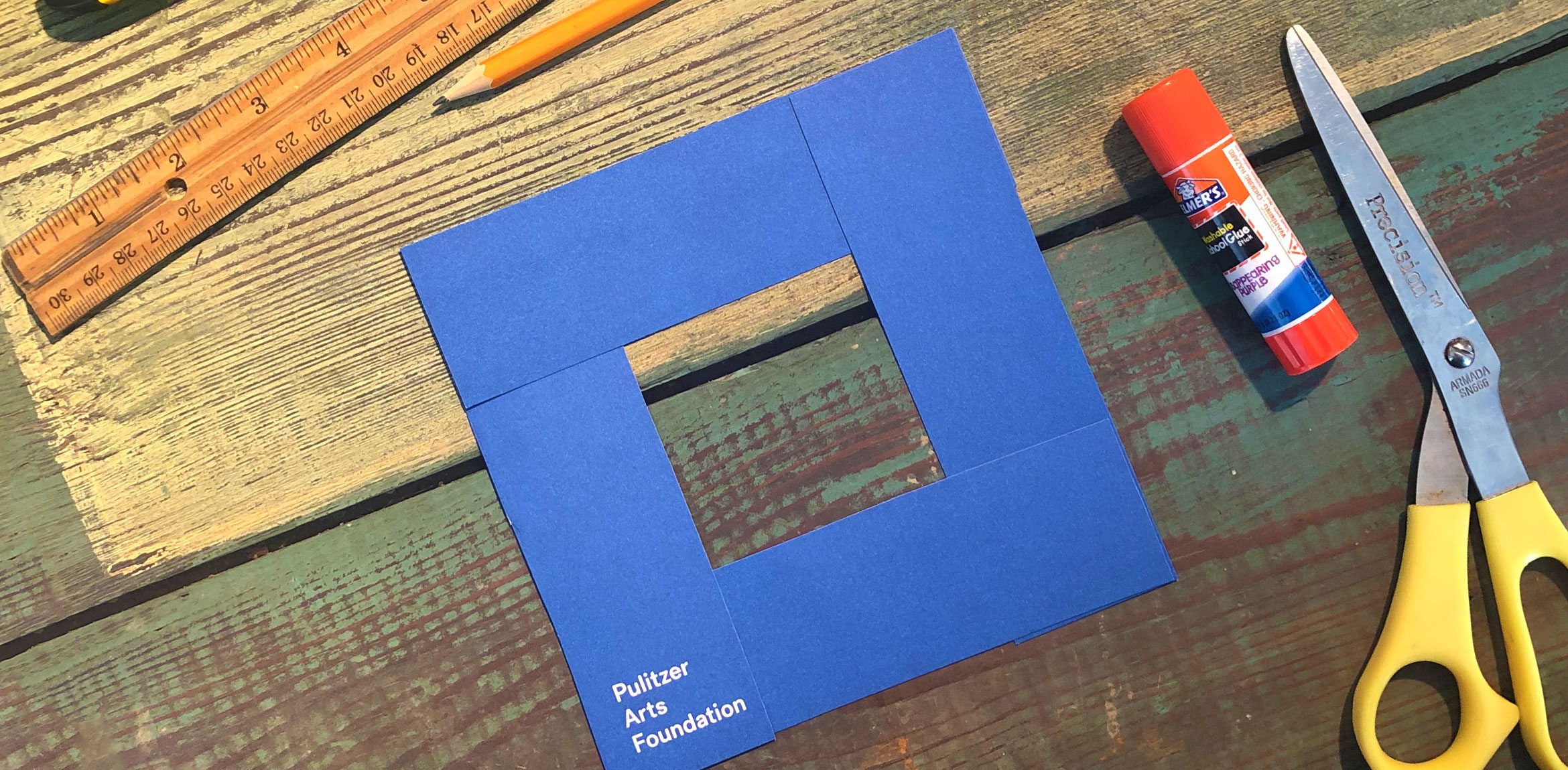A handmade square viewfinder, made with a blue Pulitzer catalogue, on a wooden surface surrounded by tools.