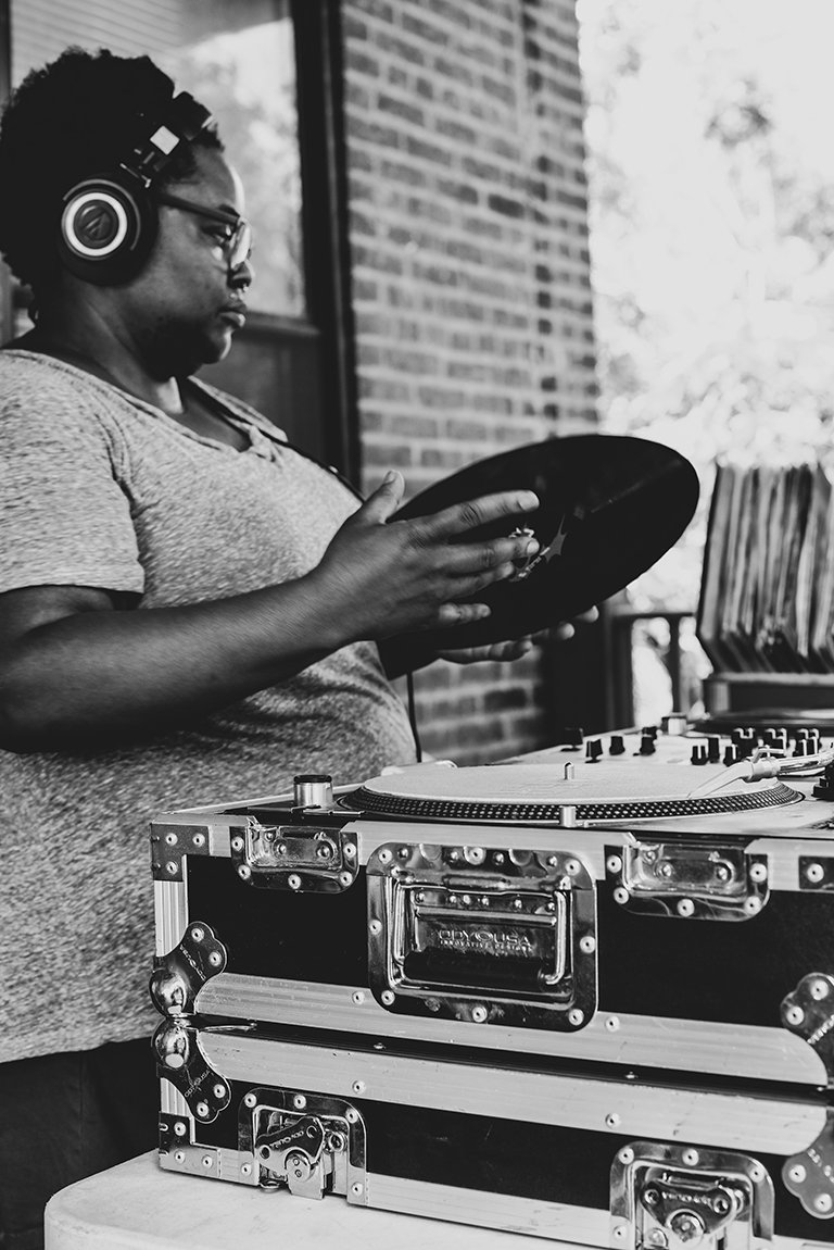 A black and white photo of Crim Dolla Cray standing behind her equipment holding a record.