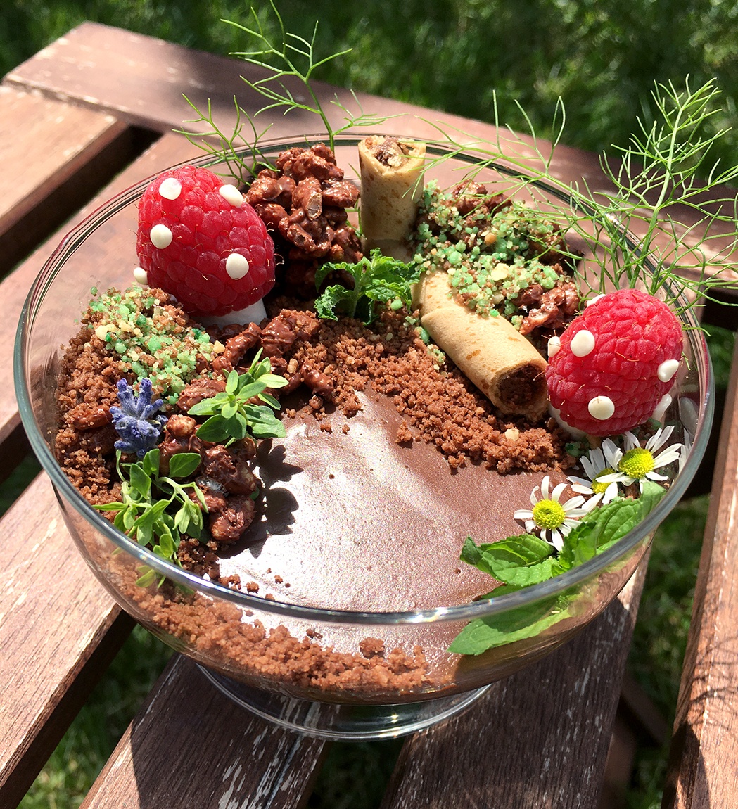 An edible arrangement in a glass bowl which includes chocolate pudding, raspberries, and flowers.