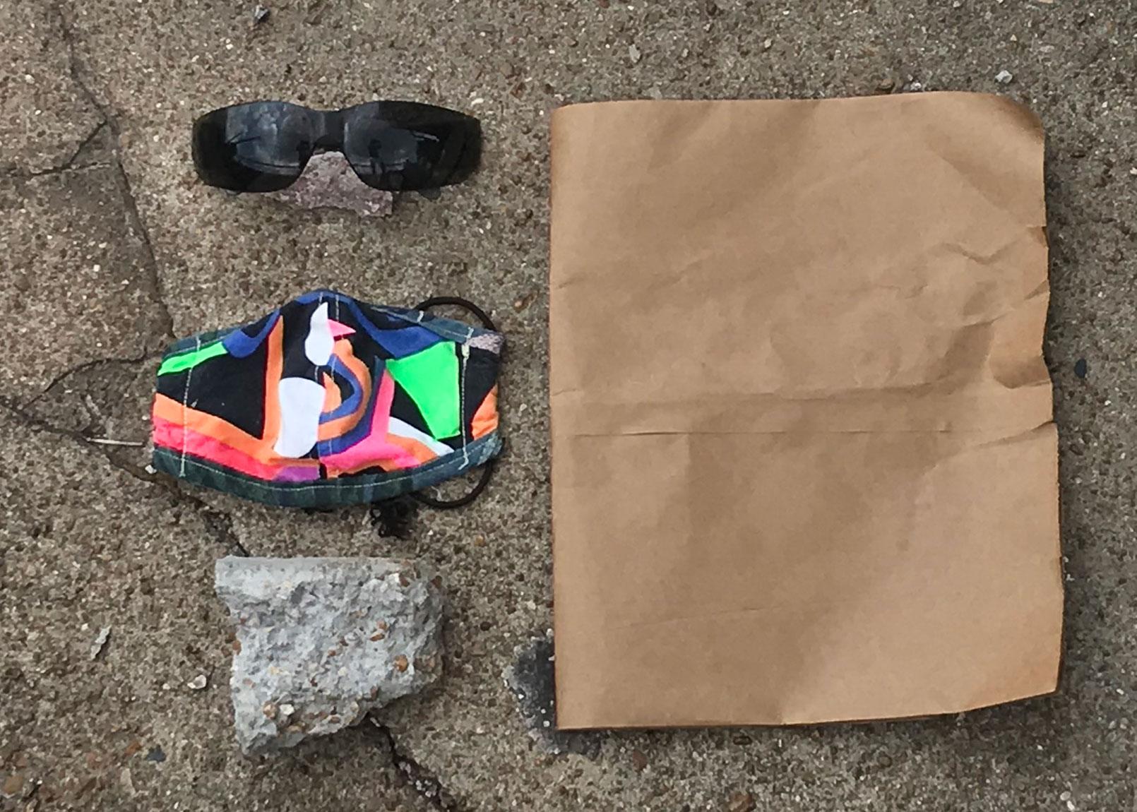 A brown piece of paper lies on the pavement beside a black pair of sunglasses, a colorful face mask, and a rock.