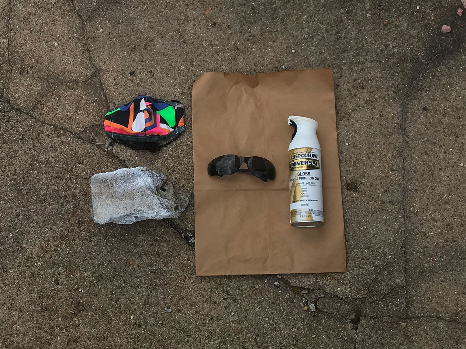 An aerial view of supplies on a pavement. A pair of glasses and a spray can of white glossy paint on a brown piece of paper and a colorful face mask and a rock beside it.