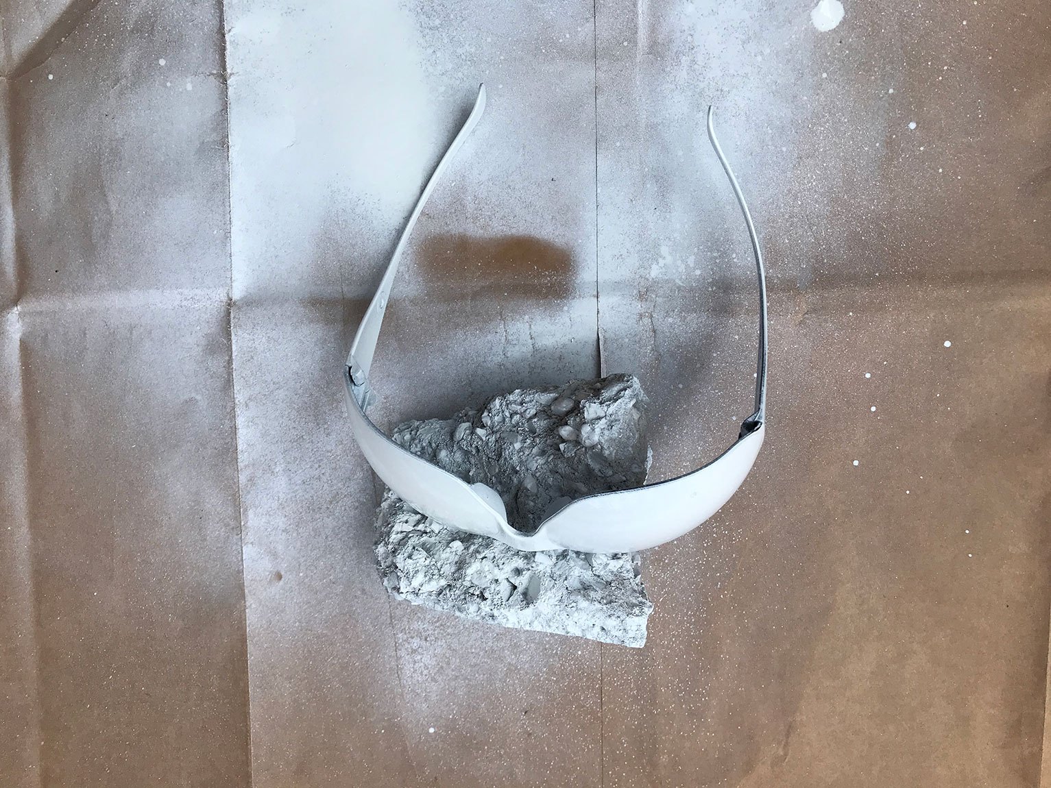 A process photo of a pair of glasses being spray painted white. They are held up by a rock on a brown piece of paper.