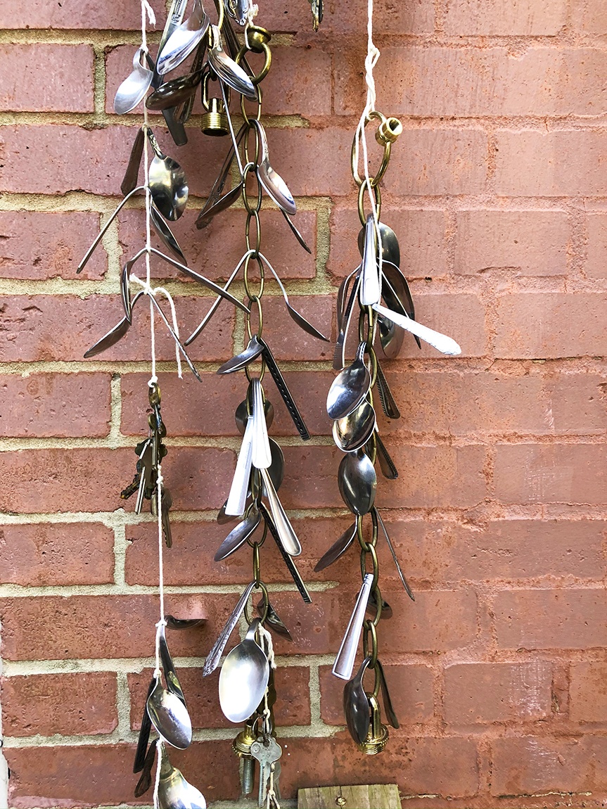 An individual holds up a metal square piece with bent spoons strung from it with string and chains in front of a brick wall.