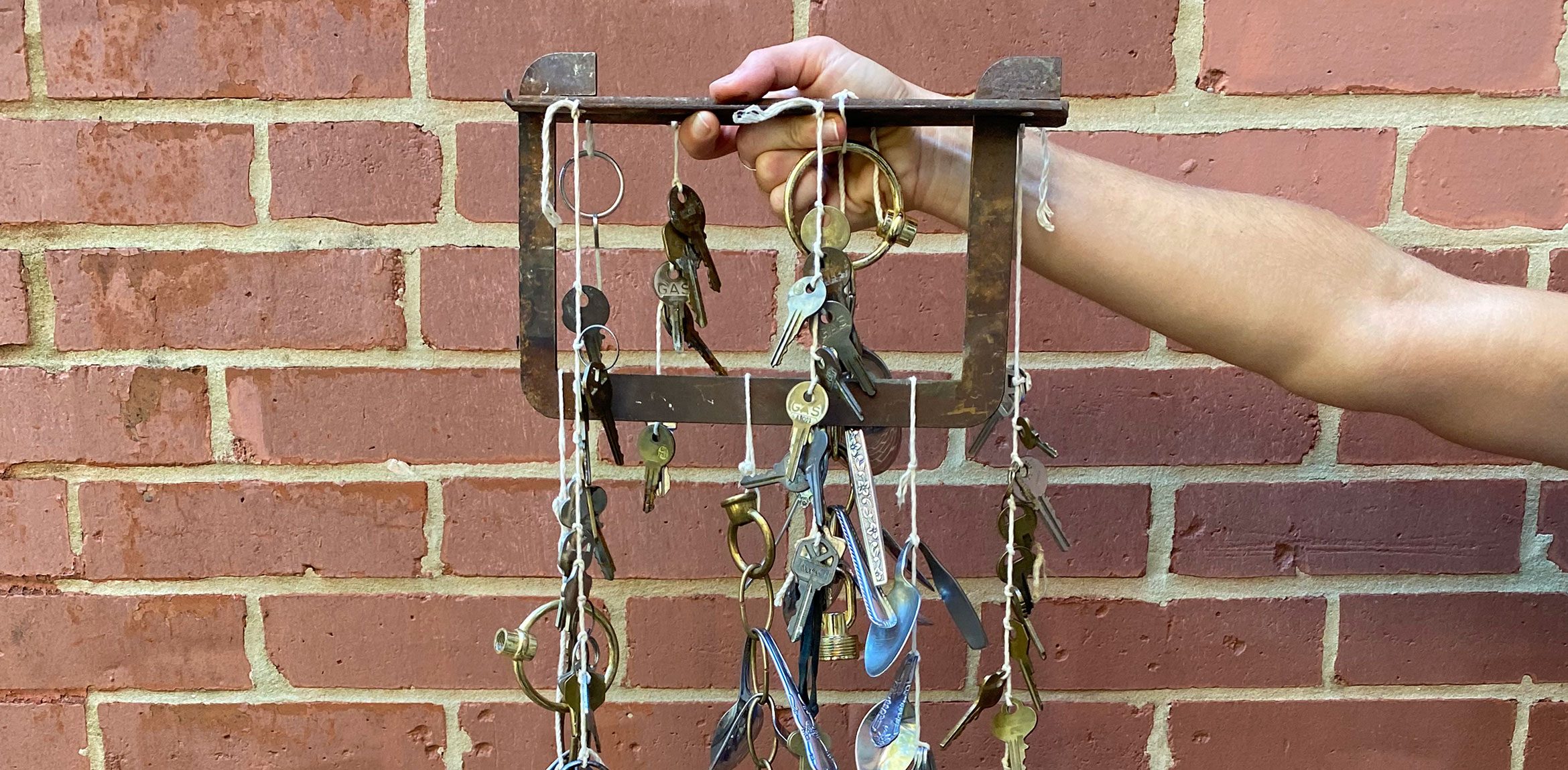 An individual holds up a metal square piece with keys and chains strung from it with string in front of a brick wall.