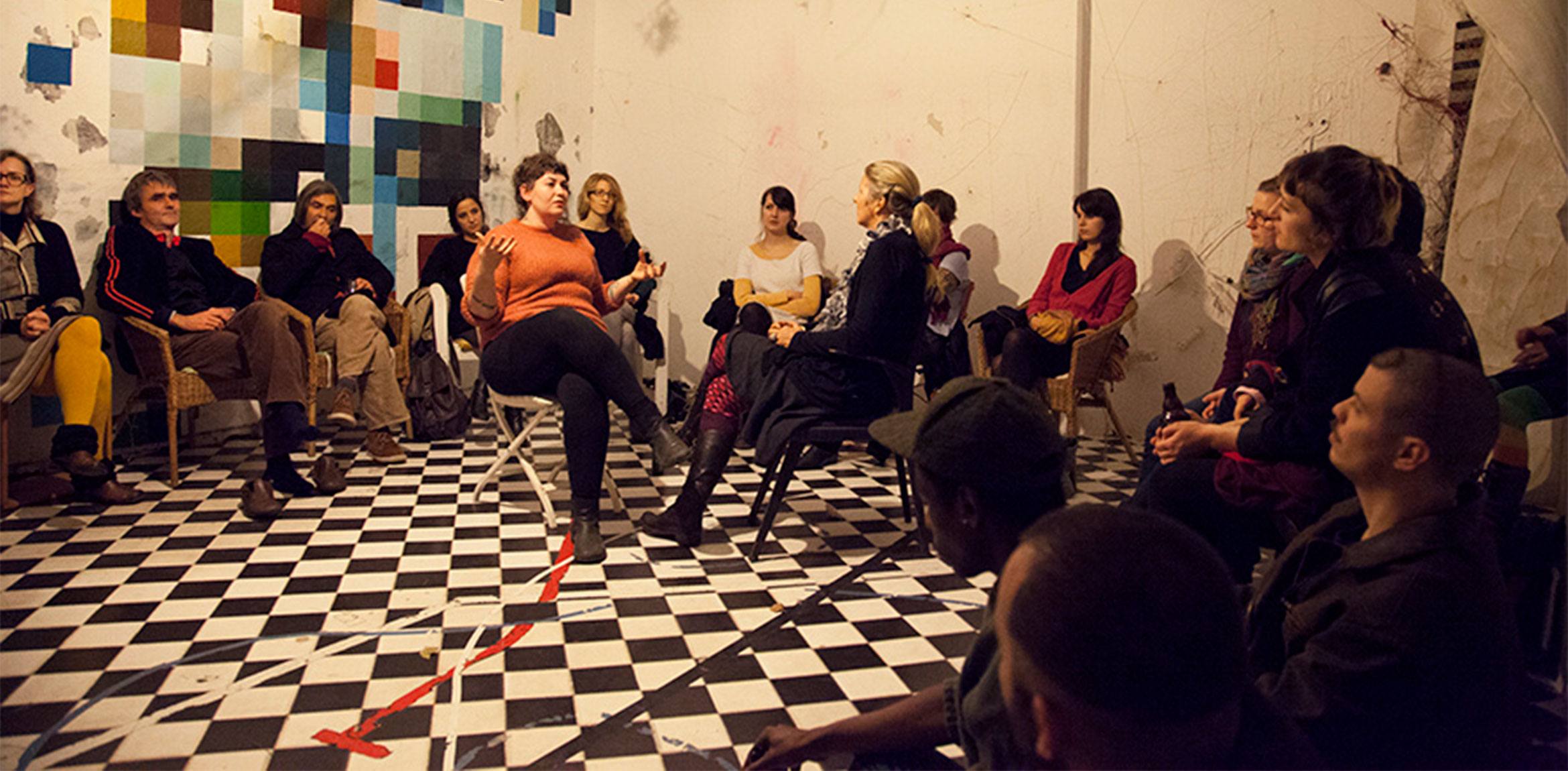 Lindsay Tunkl and Aja Bond sit and converse in the center of a circle of seated guests on a checkboard floor.