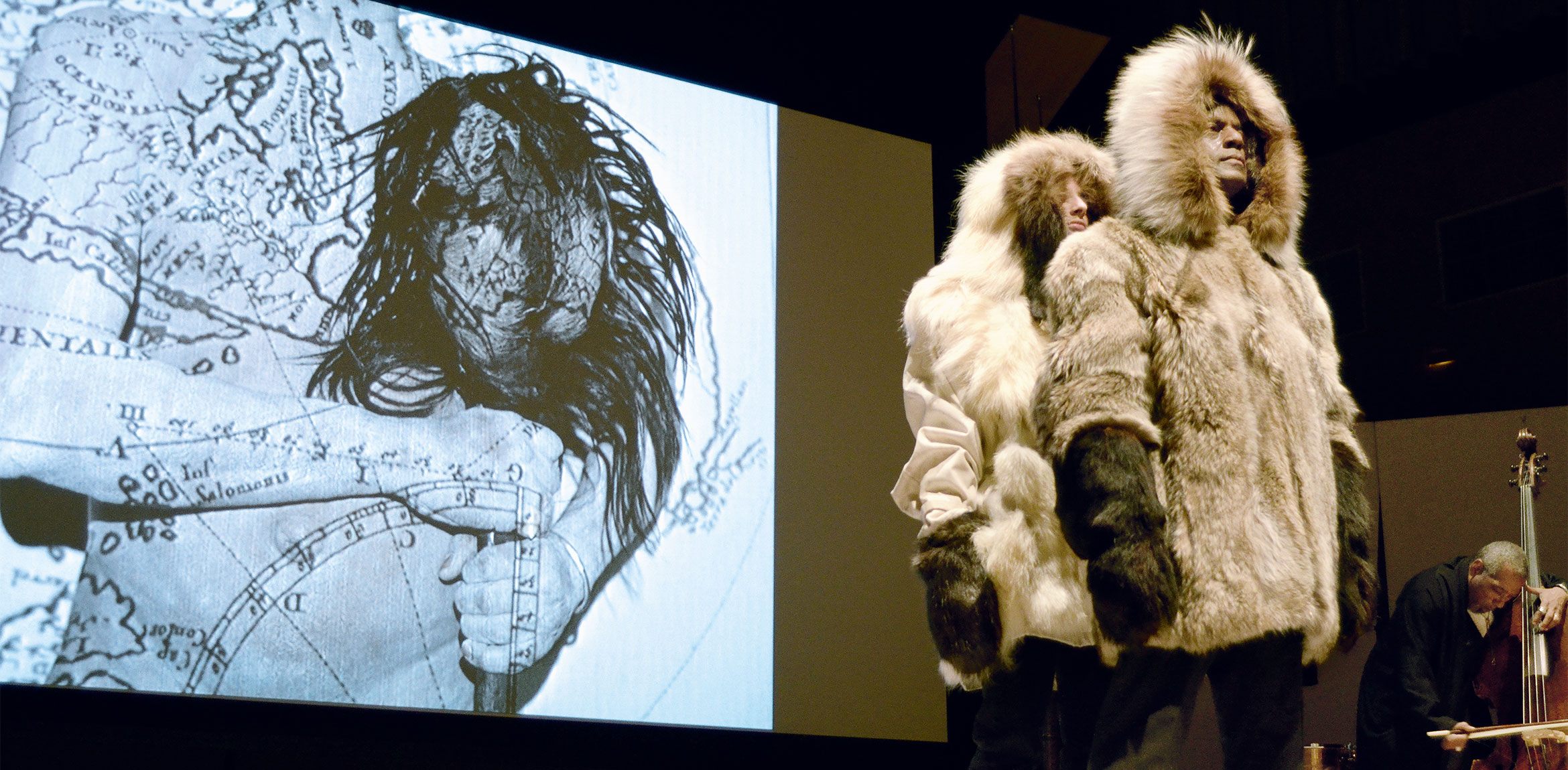 Two Lone Wolf Recital Corps members wear Alaskan fur parkas and stand on a stage in front of an image projected on a screen behind them. A cellist plays to the right.