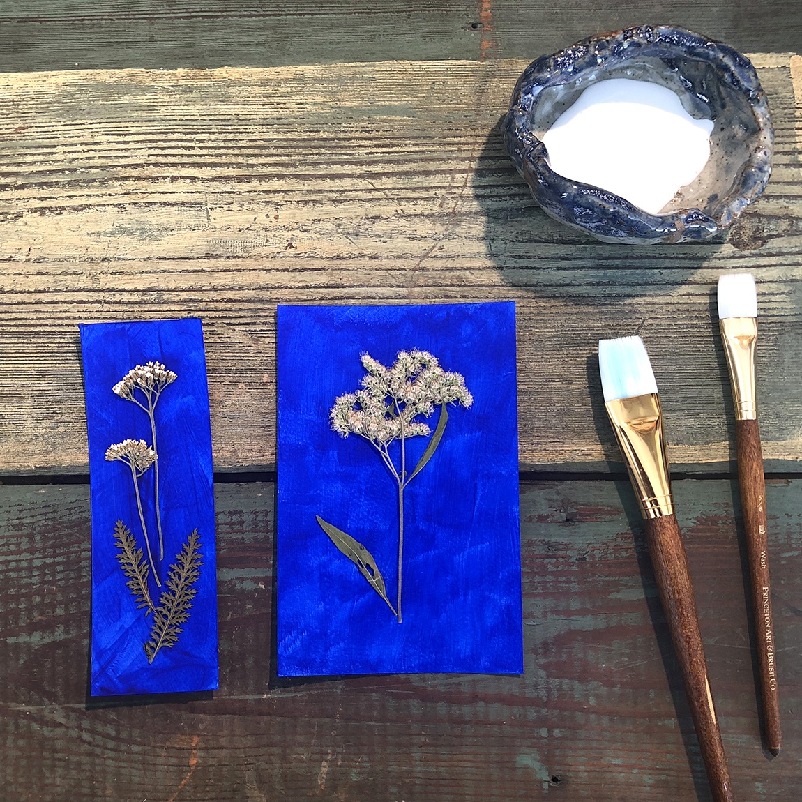 Two ultramarine paper bookmarks with plants arranged on them, beside two flat brushes and a container holding glue.