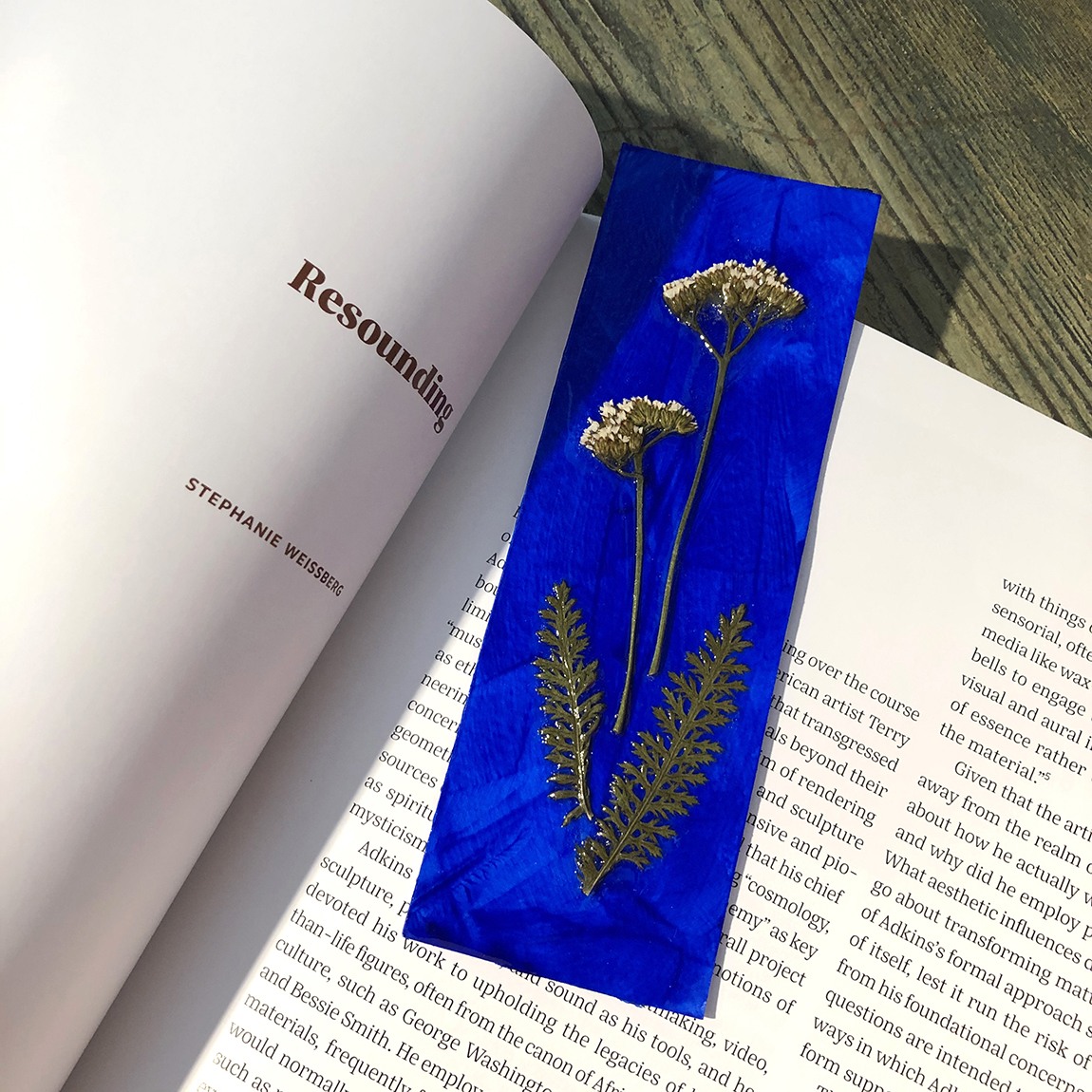 Four pressed plants sealed to an ultramarine paper bookmark, sitting on a page of the exhibition catalogue for "Terry Adkins: Resounding."