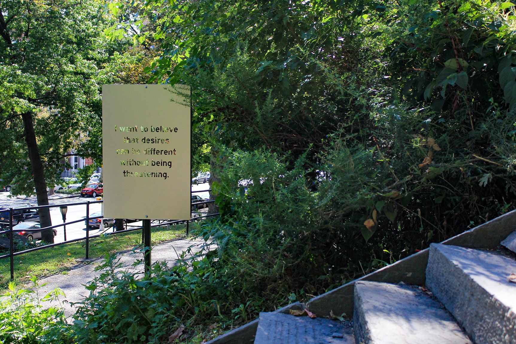 Chloë Bass' mirrored lettering piece, "I want to believe that desires can be different without being threatening. The part of you that says 'I can share myself with strangers.'" The piece is installed beside a sidewalk, surrounded by trees.