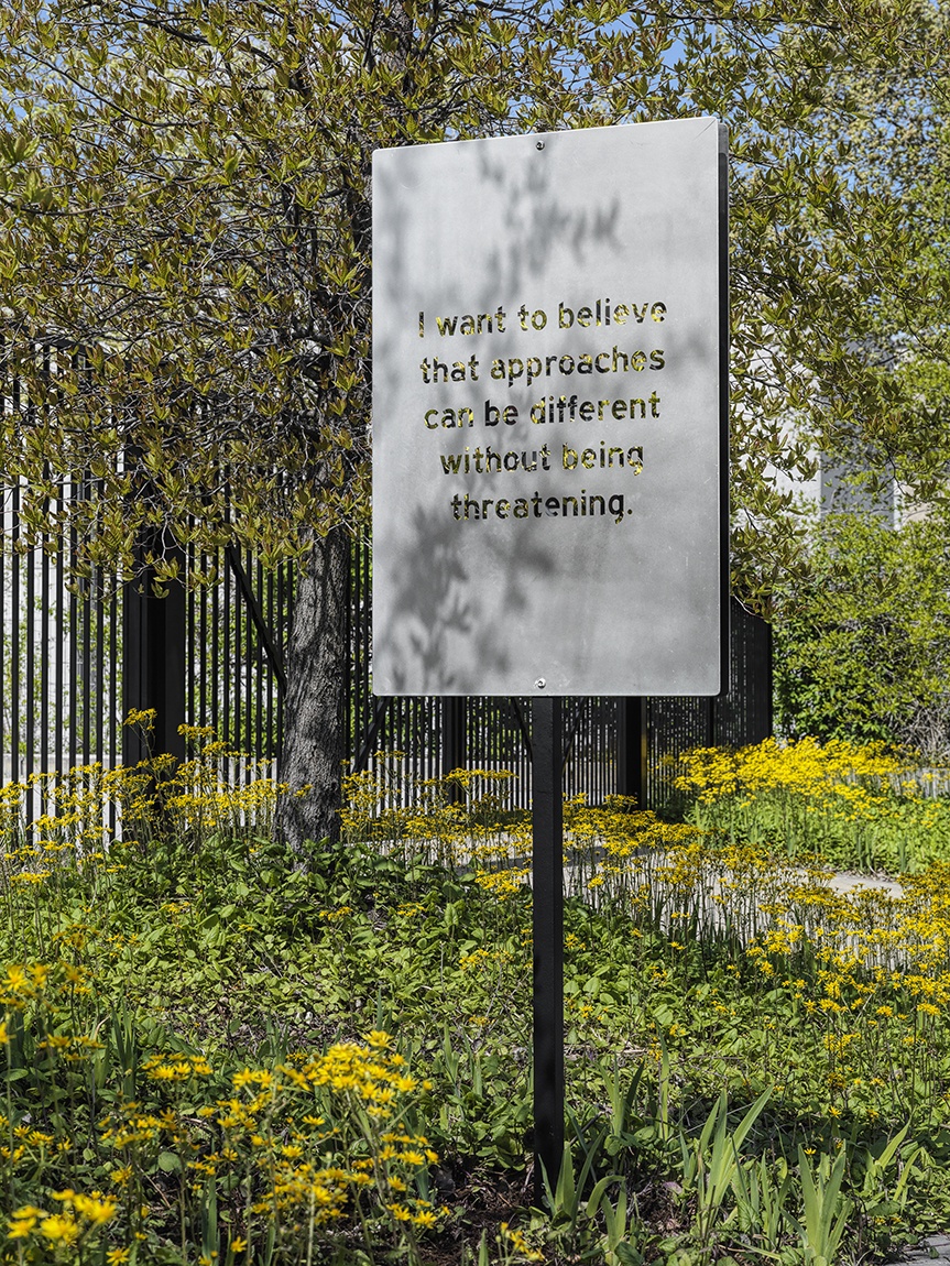An aluminum sign artwork with text by Chloë Bass installed amongst yellow flowers