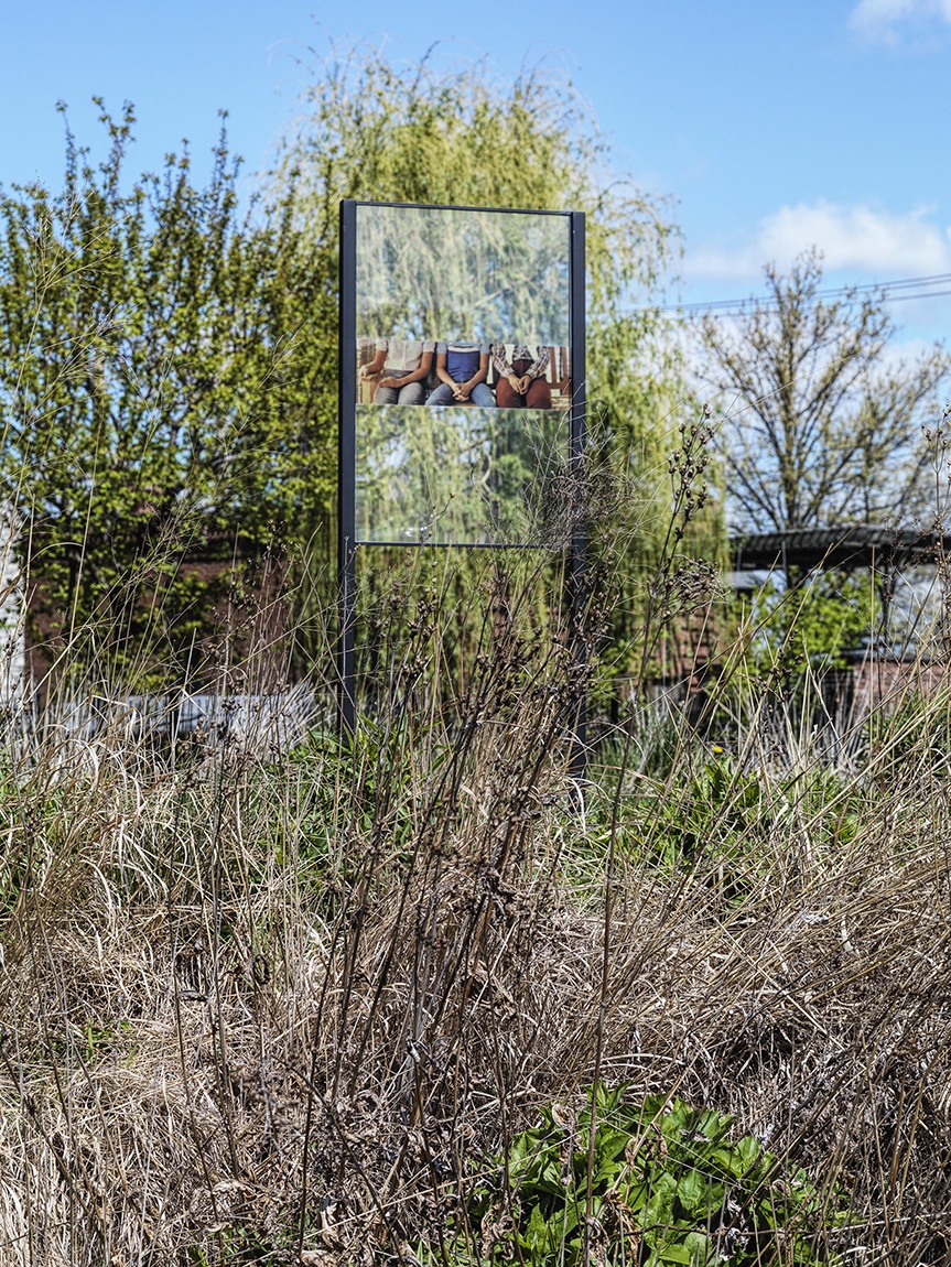 An acrylic sign artwork by Chloë Bass featuring a cropped image of three people sitting, installed outdoors
