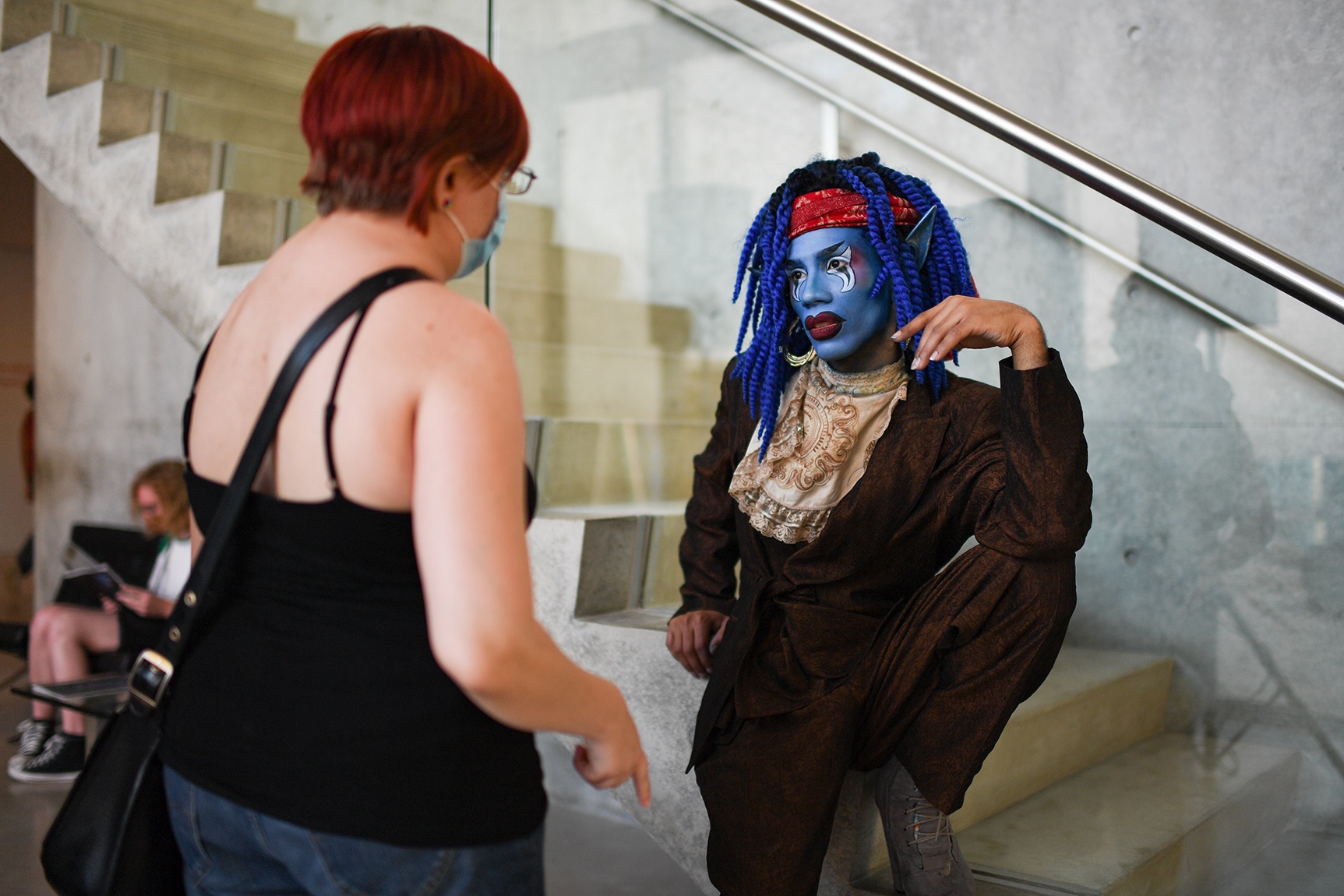 Maxi Glamour wearing blue facial makeup sits perched on the mezzanine stairs while talking to a program visitor