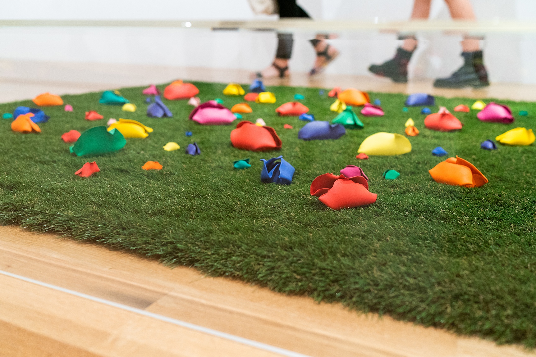 Brightly colored clay forms place on artificial turf, beneath a vitrine. Two pairs of legs are in the background.