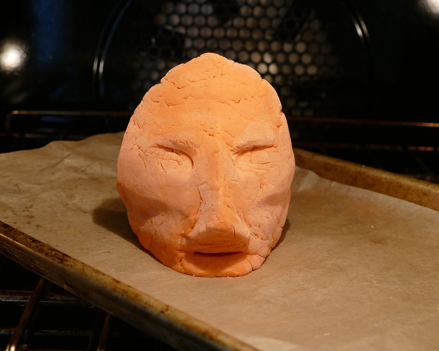 A head formed from handmade clay, on a baking sheet in an oven.