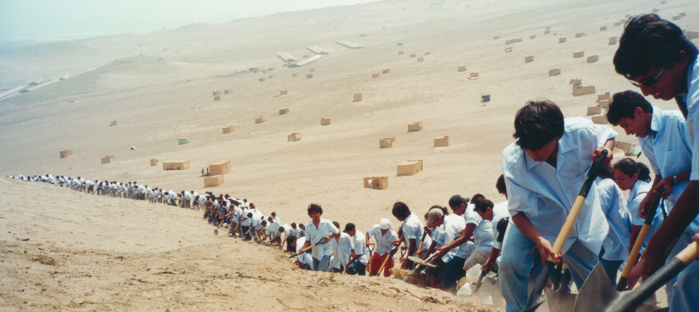 A line of hundreds of participants in artist Francis Alys's "When Faith Moves Mountains." They are assembled on a sand dune with shovels in hand