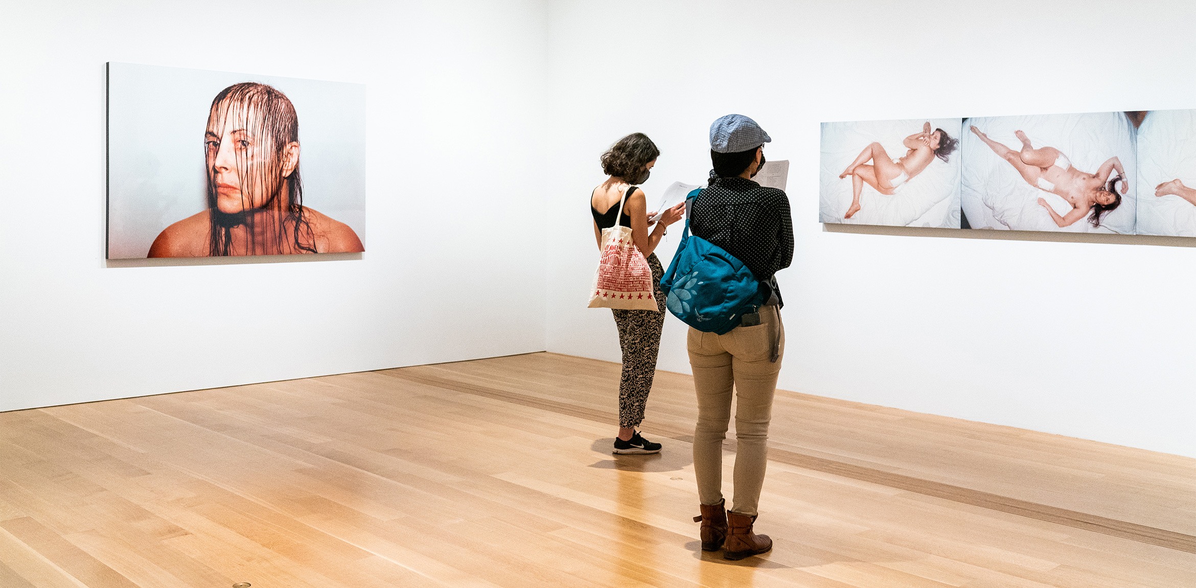 Two visitors standing in the East Gallery. On the left is a photograph portrait of the artist Hannah Wilke and on the right is a triptych featuring views of Hannah Wilke from above.