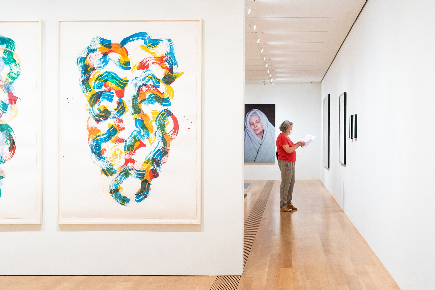 A visitor stands in the galleries; in the foreground is a large colorful abstract portrait of a face; in the background, a portion of a self-portrait is visible