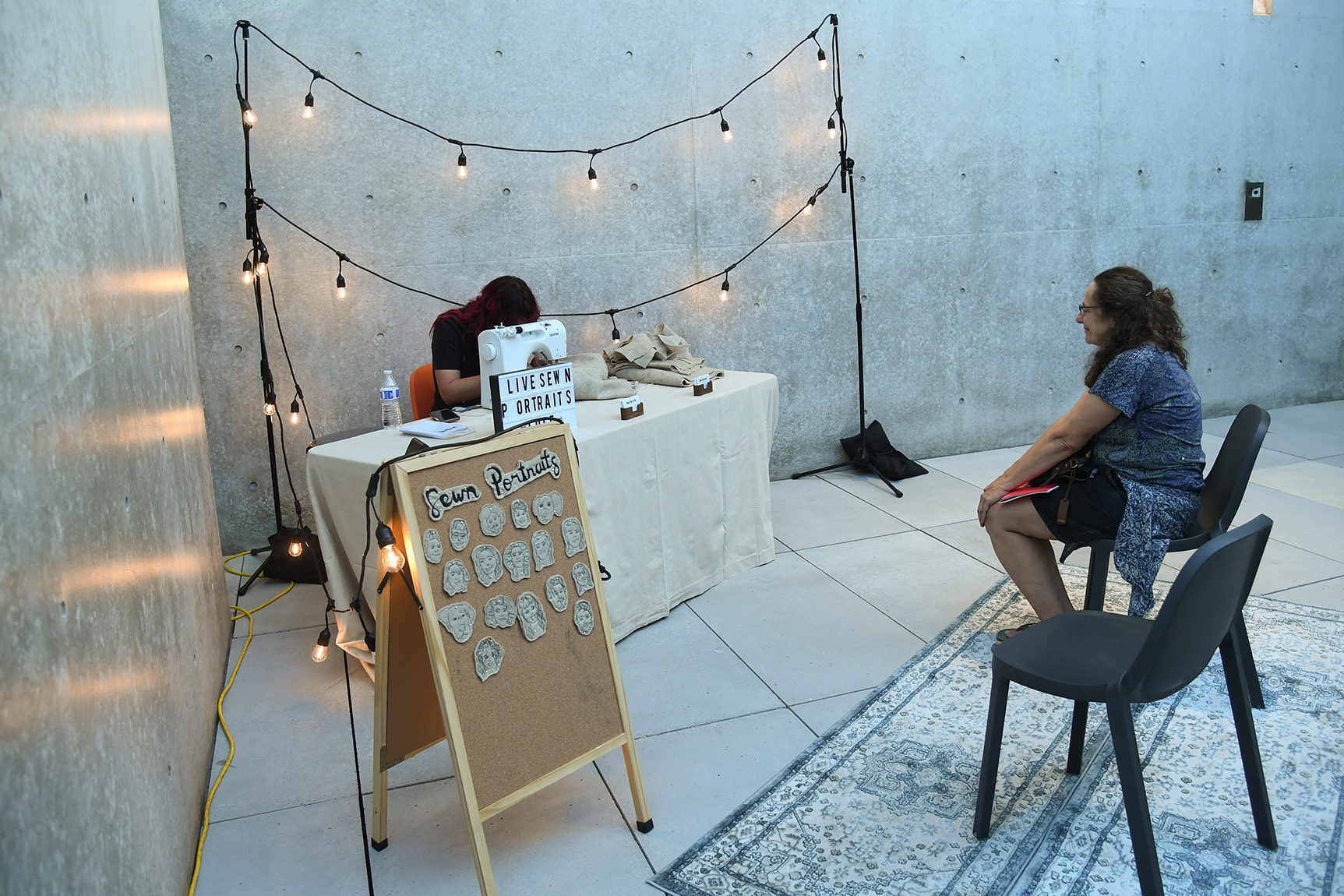 Artist Tucker Pierce working at a sewing machine, a seated visitor, and a corkboard with several sewn portrait examples