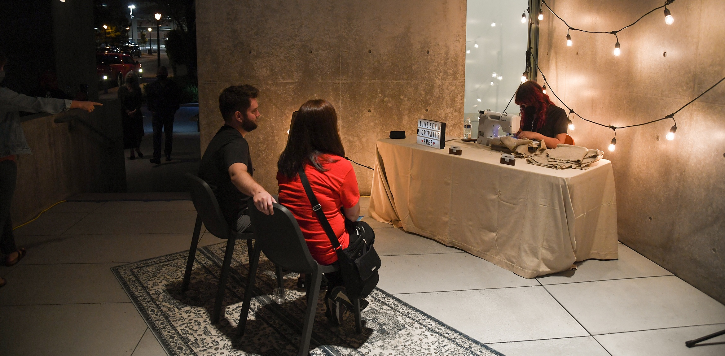 Artist Tucker Pierce sewing at a table with two visitors sitting for their portrait