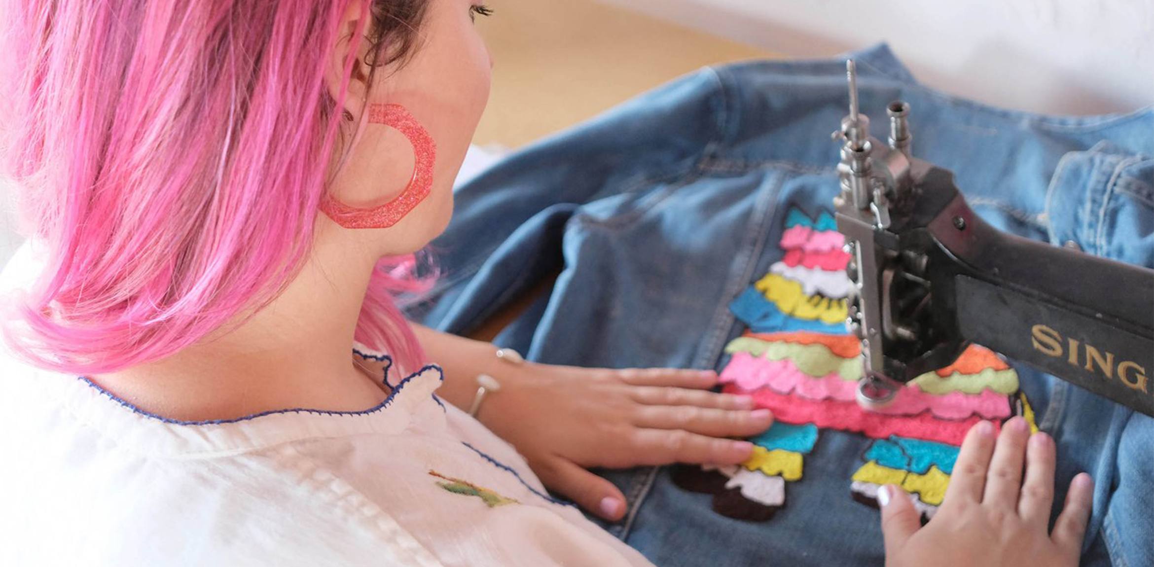 An individual with pink hair embroidering a denim jacket with colorful threads