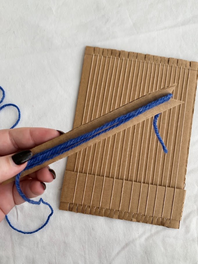 A strung cardboard loom and a hand holding a cardboard shuttle, wrapped with blue yarn