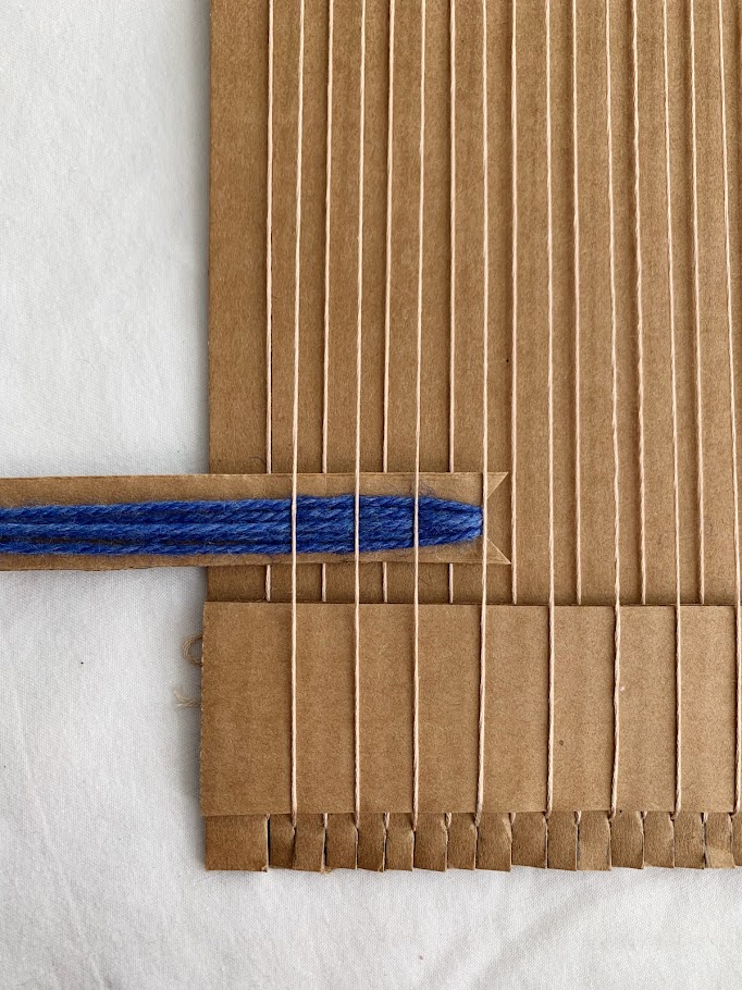 A cardboard shuttle wrapped with yarn that is partially inserted into a strung cardboard loom
