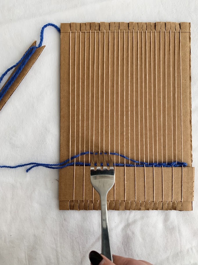 A fork pulling down woven yarn in a cardboard loom